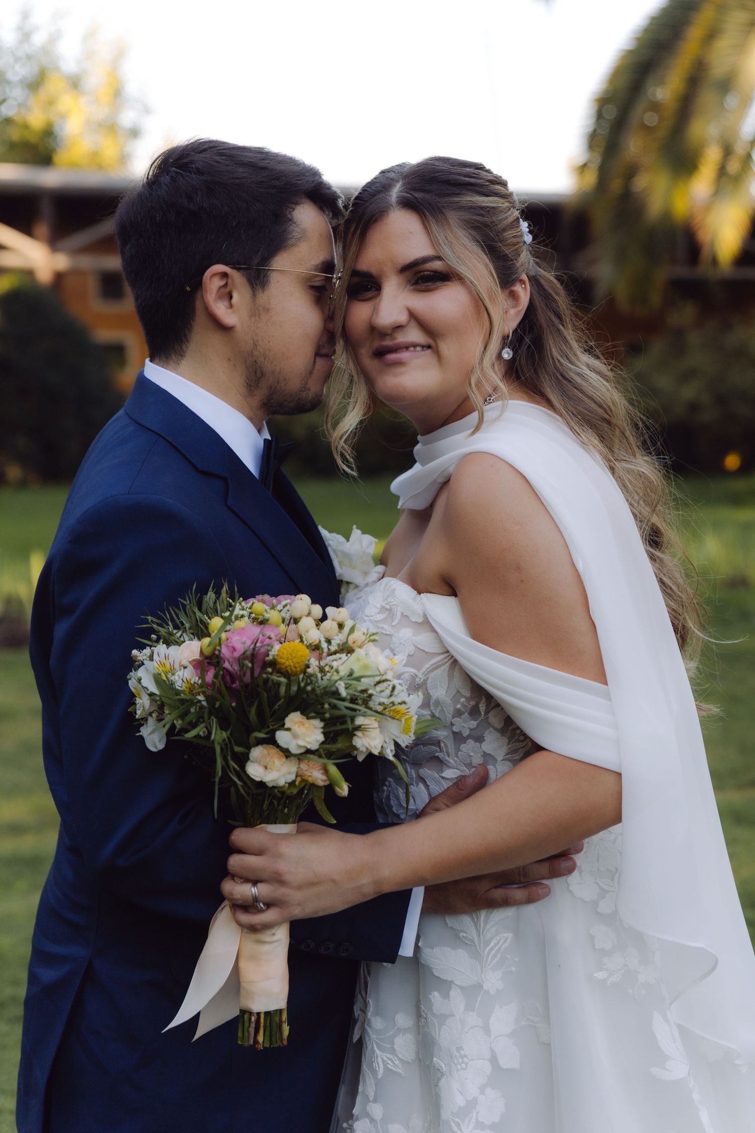 Groom tenderly kisses bride while she smiles holding a colorful spring wedding bouquet outdoors.