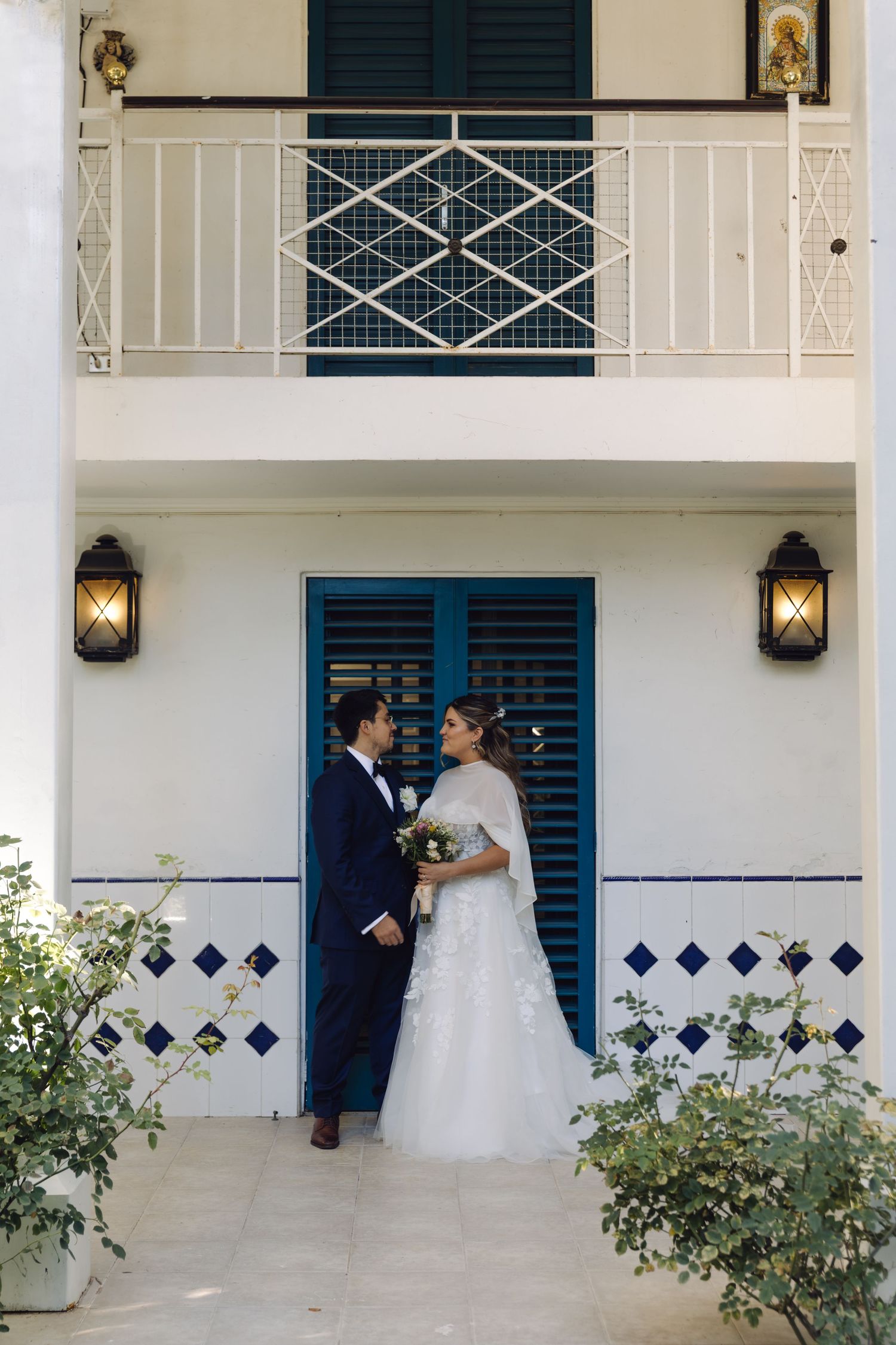 Bride and groom pose romantically in front of white building with blue shutters and decorative tile accents.