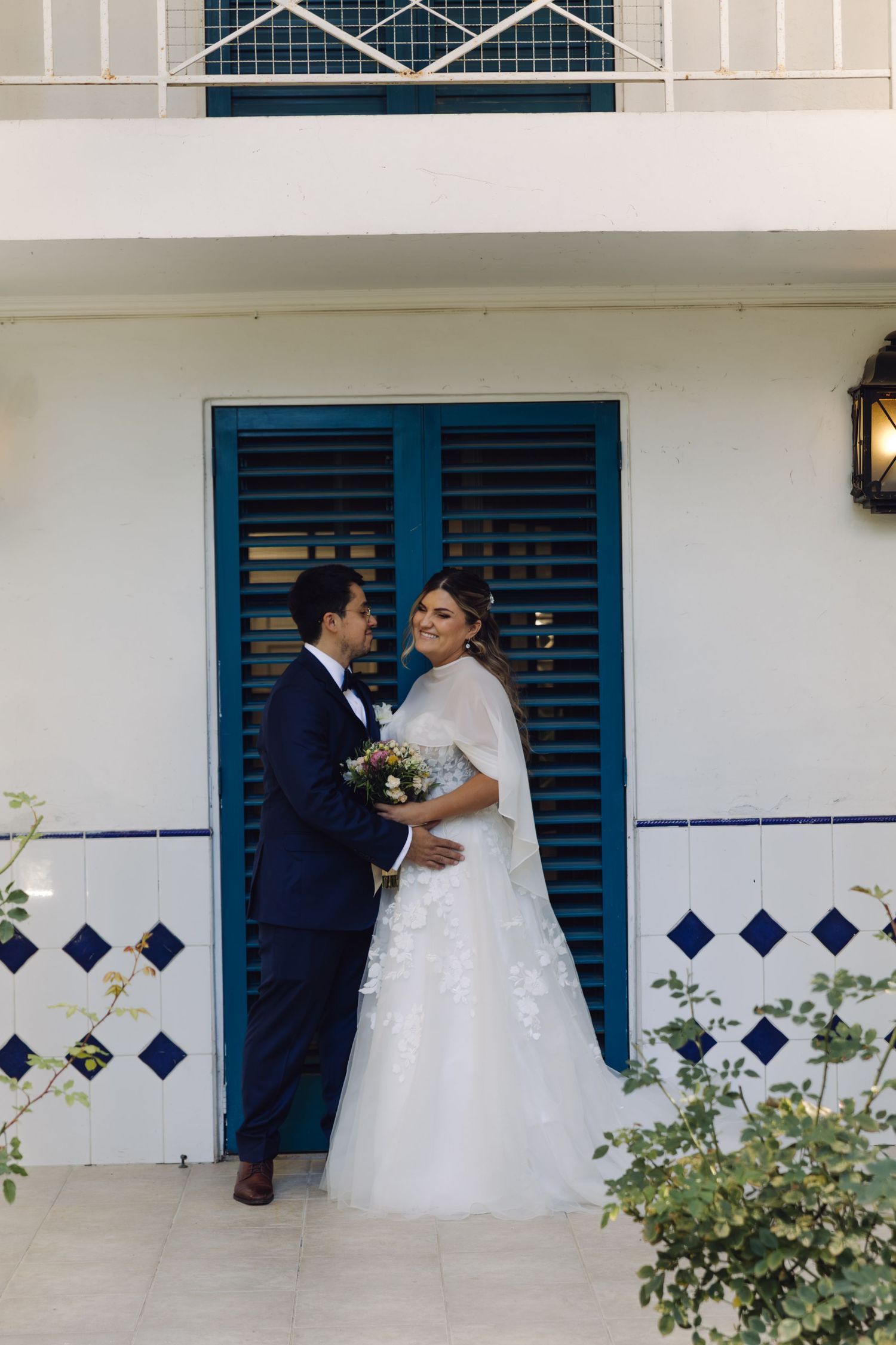 Bride and groom share an intimate moment leaning against white wall with blue shuttered doors behind them.