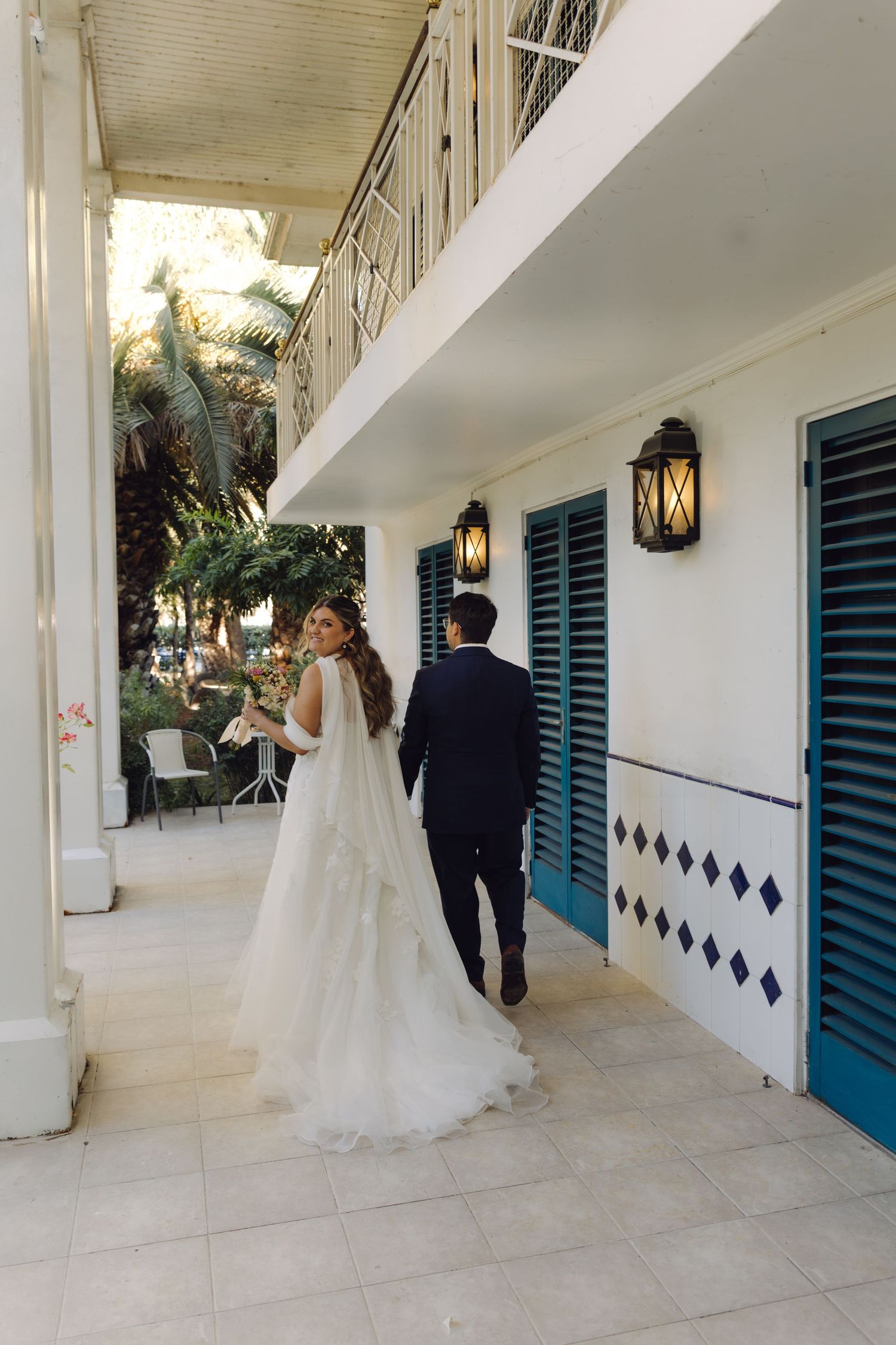 Bride looks back smiling as couple walks hand in hand along a covered veranda with blue shuttered doors.