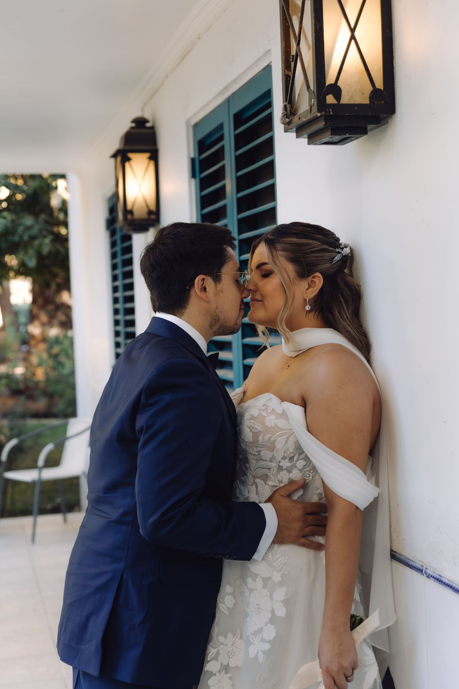 Bride and groom share a romantic kiss against white wall flanked by vintage lanterns and blue shutters.