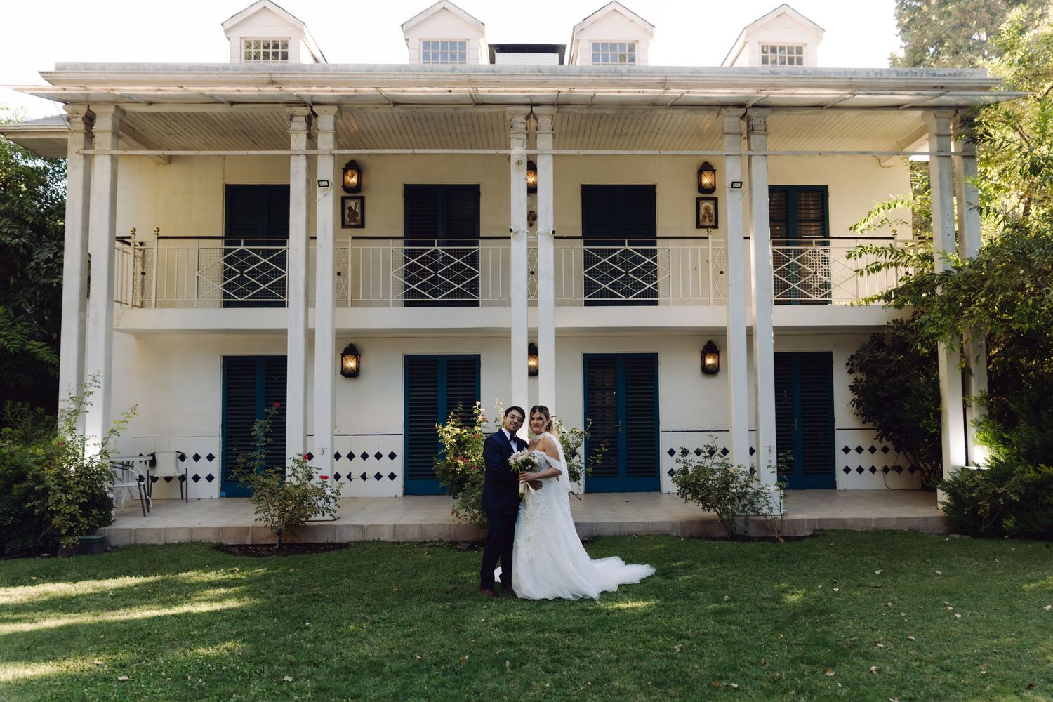 Bride and groom pose together on lawn in front of elegant two-story white colonial mansion with blue shutters.