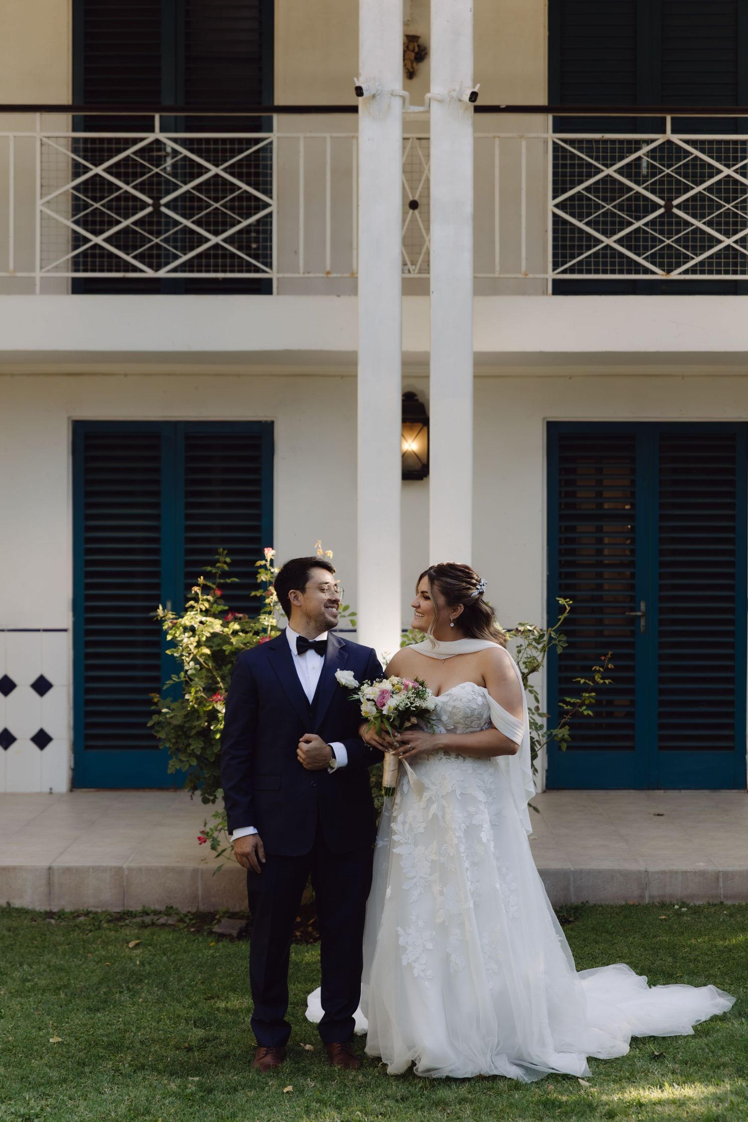 Bride and groom share a romantic moment in front of a white colonial building with teal shutters.