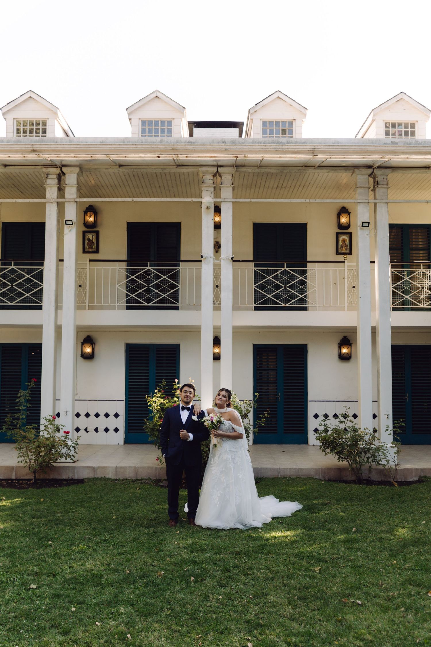 Wedding couple poses on green lawn before elegant white two-story colonial venue with blue shutters.