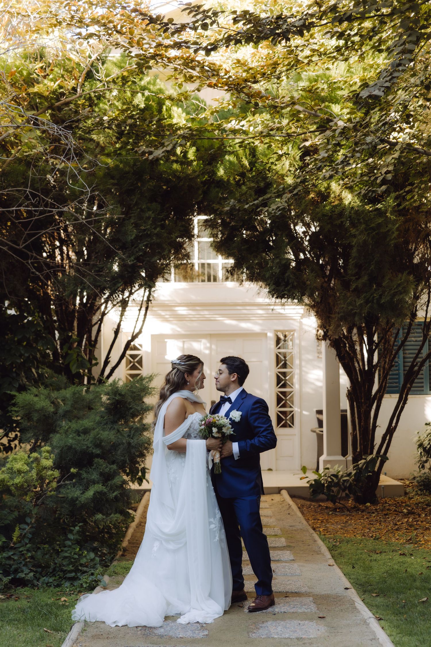 Bride and groom share an intimate moment on a garden path framed by lush green trees.