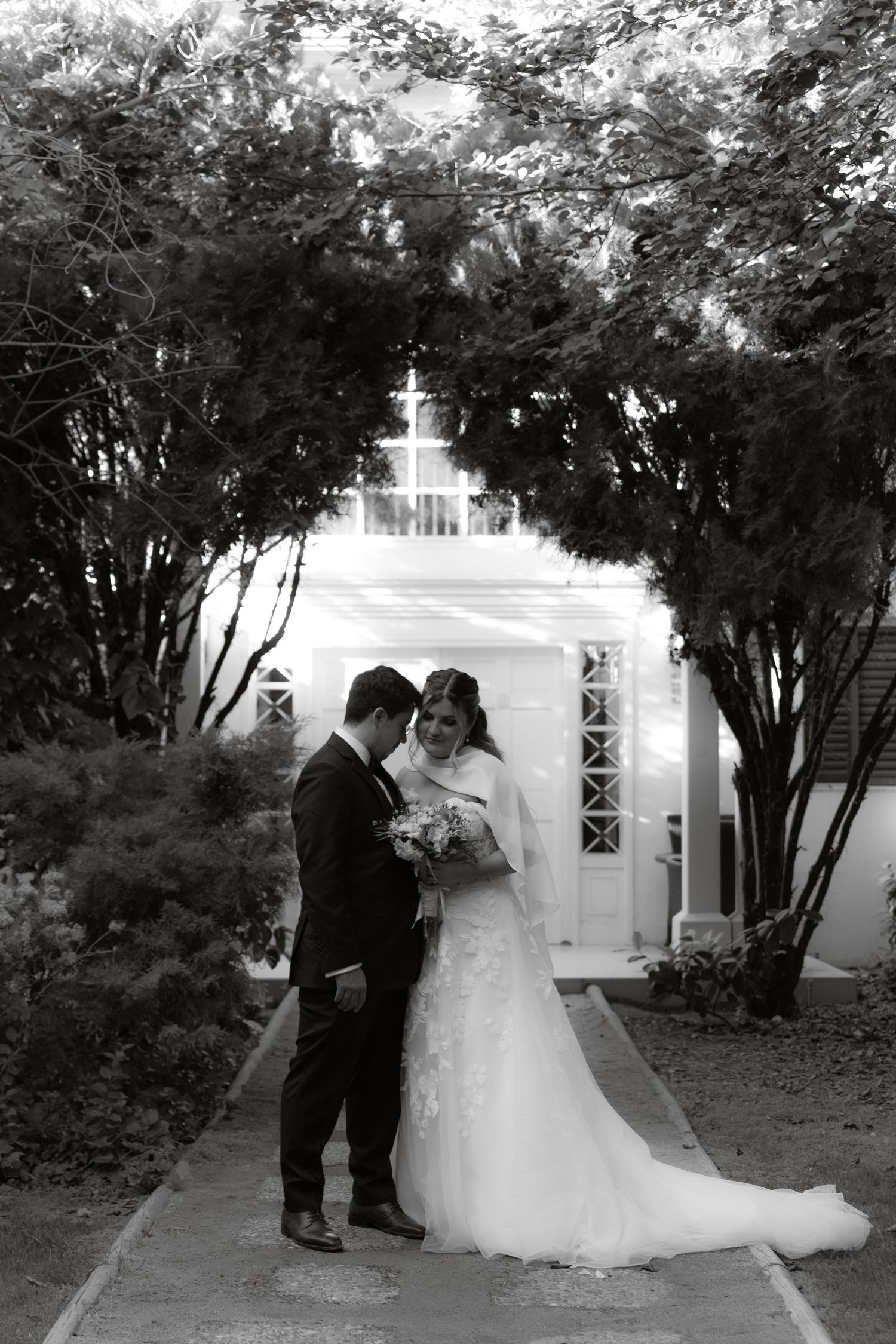 Black and white photo of bride and groom embracing on garden path before white chapel entrance.