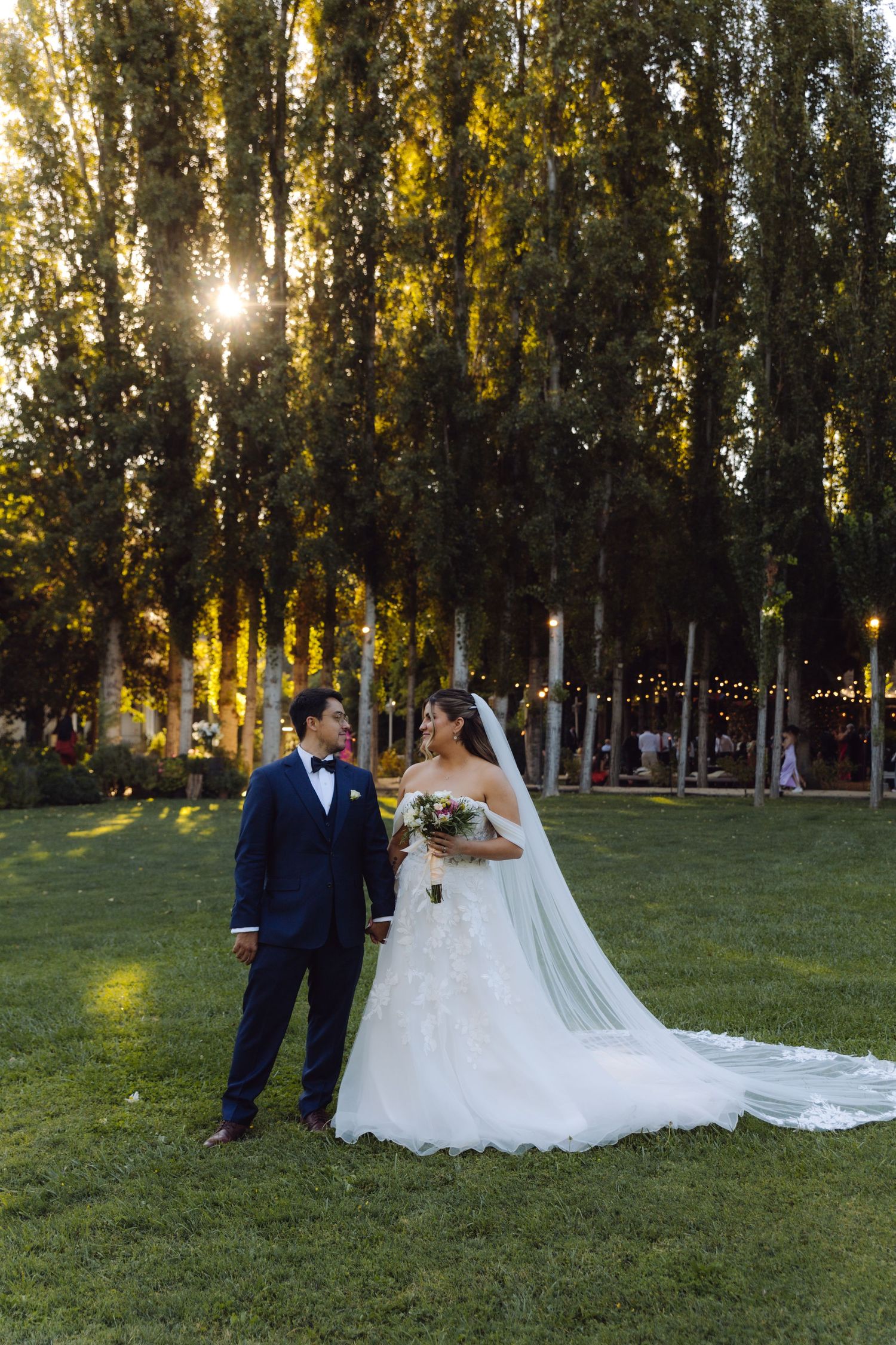 Bride and groom walk through sunlit garden with tall trees and string lights at golden hour.