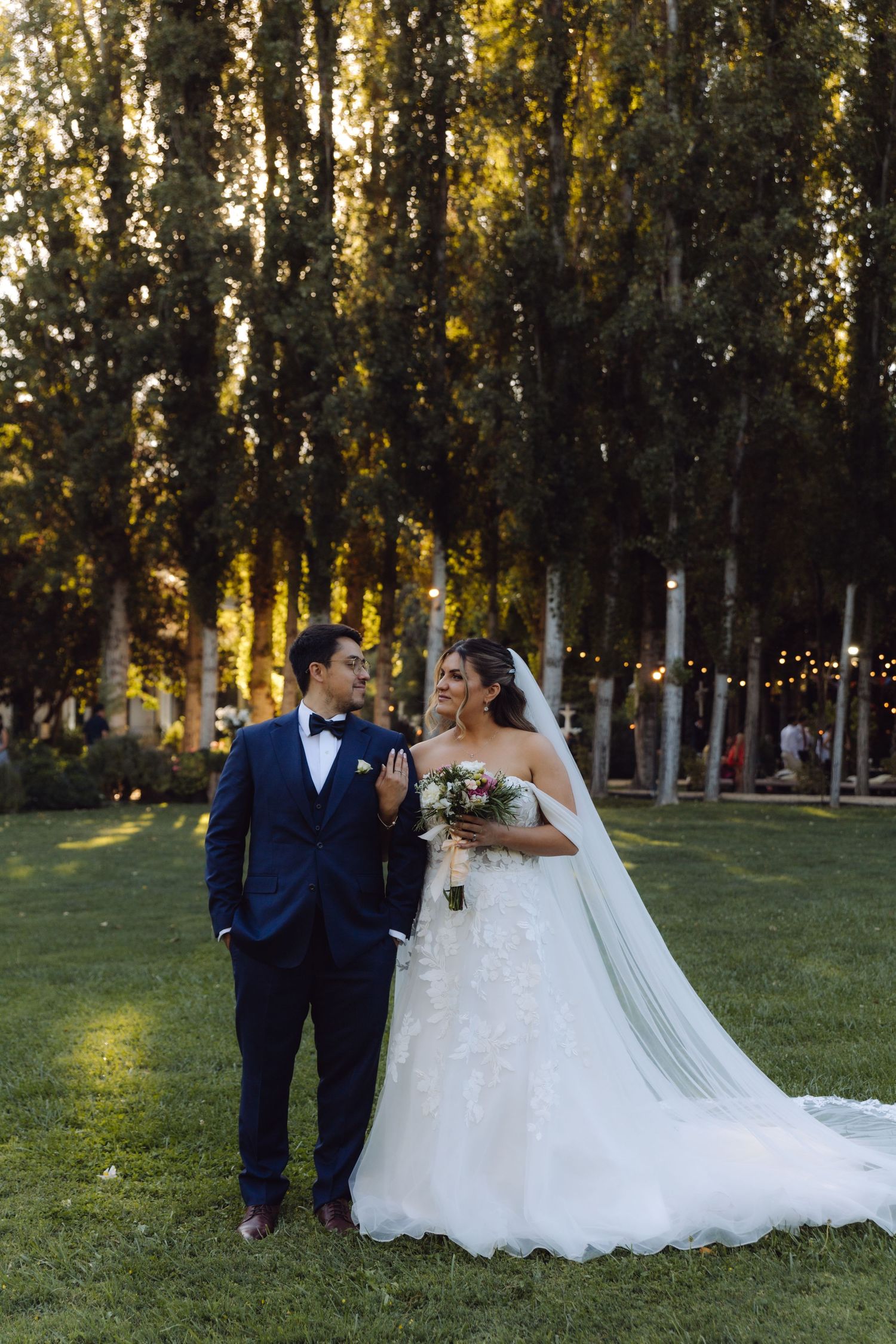 Elegant couple shares a loving glance on green lawn surrounded by tall trees at sunset.