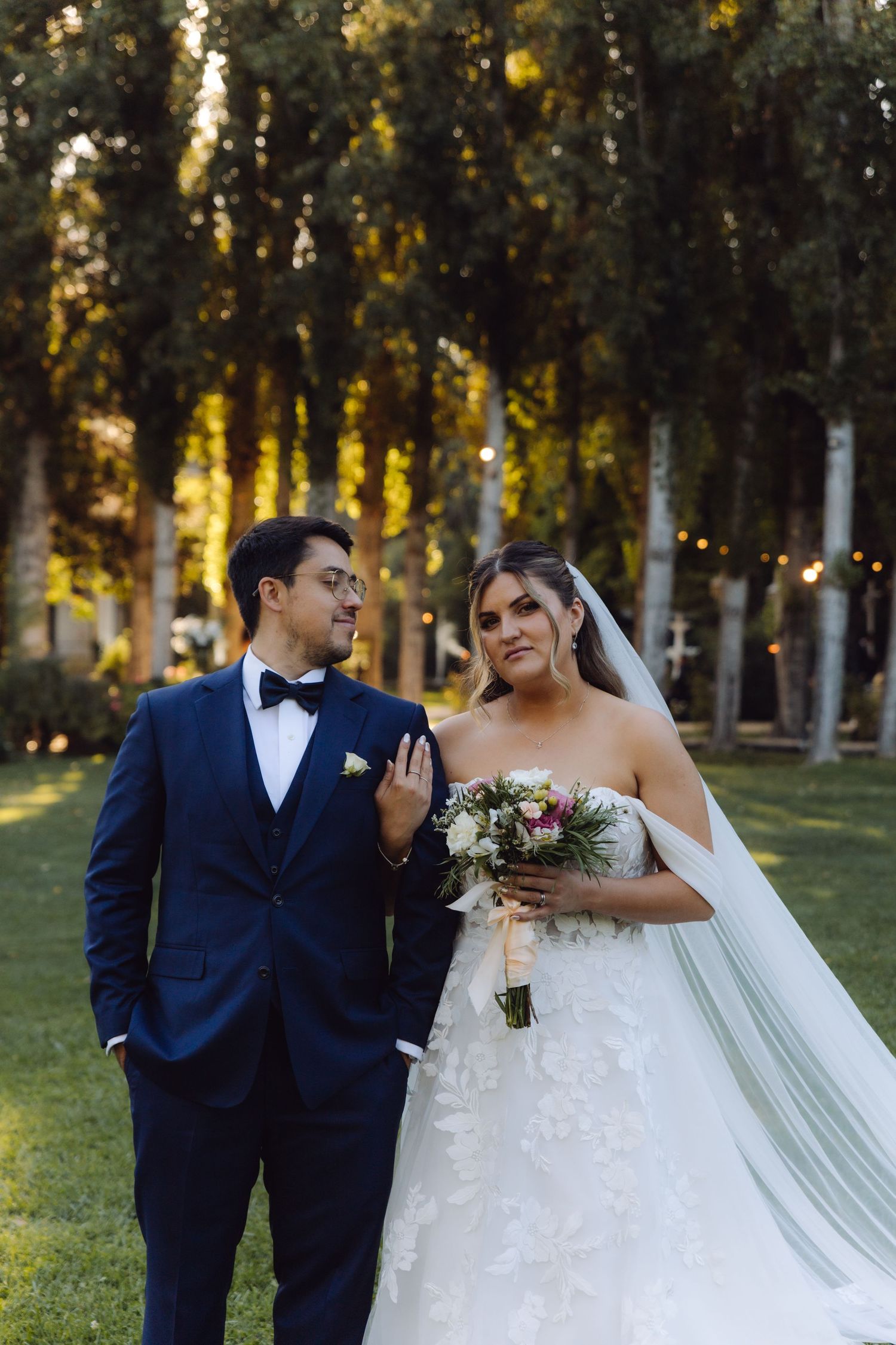 Groom in navy tuxedo and bride in lace gown walk together in sunlit garden setting.
