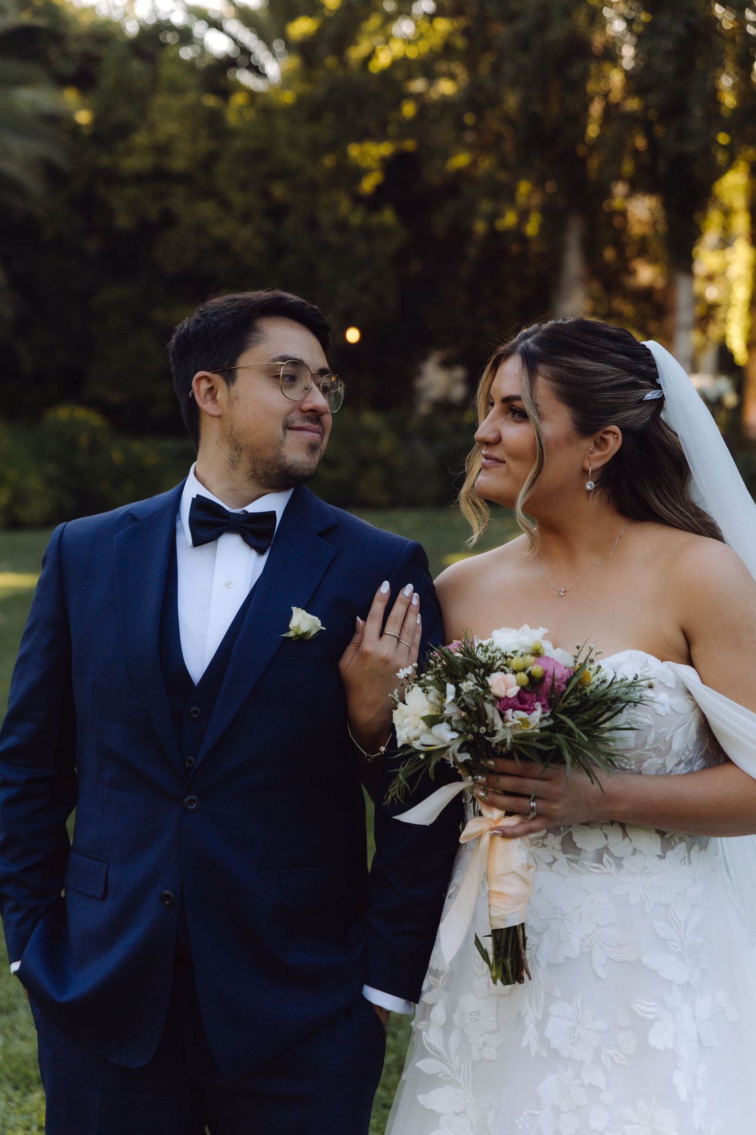 Smiling bride and groom share a tender look outdoors in a lush garden at dusk.