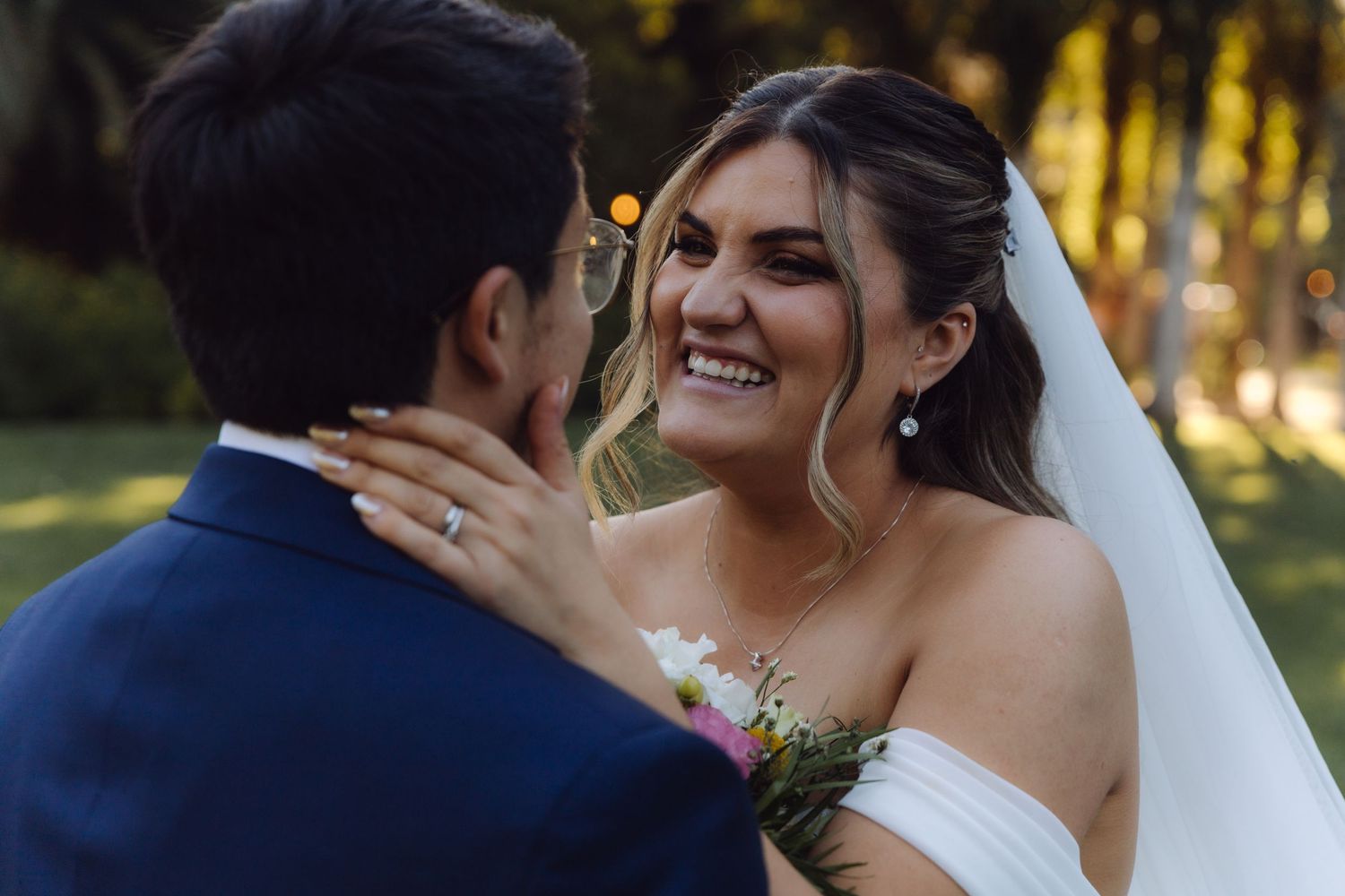 Radiant bride smiles joyfully at groom while holding a floral bouquet in an outdoor garden.