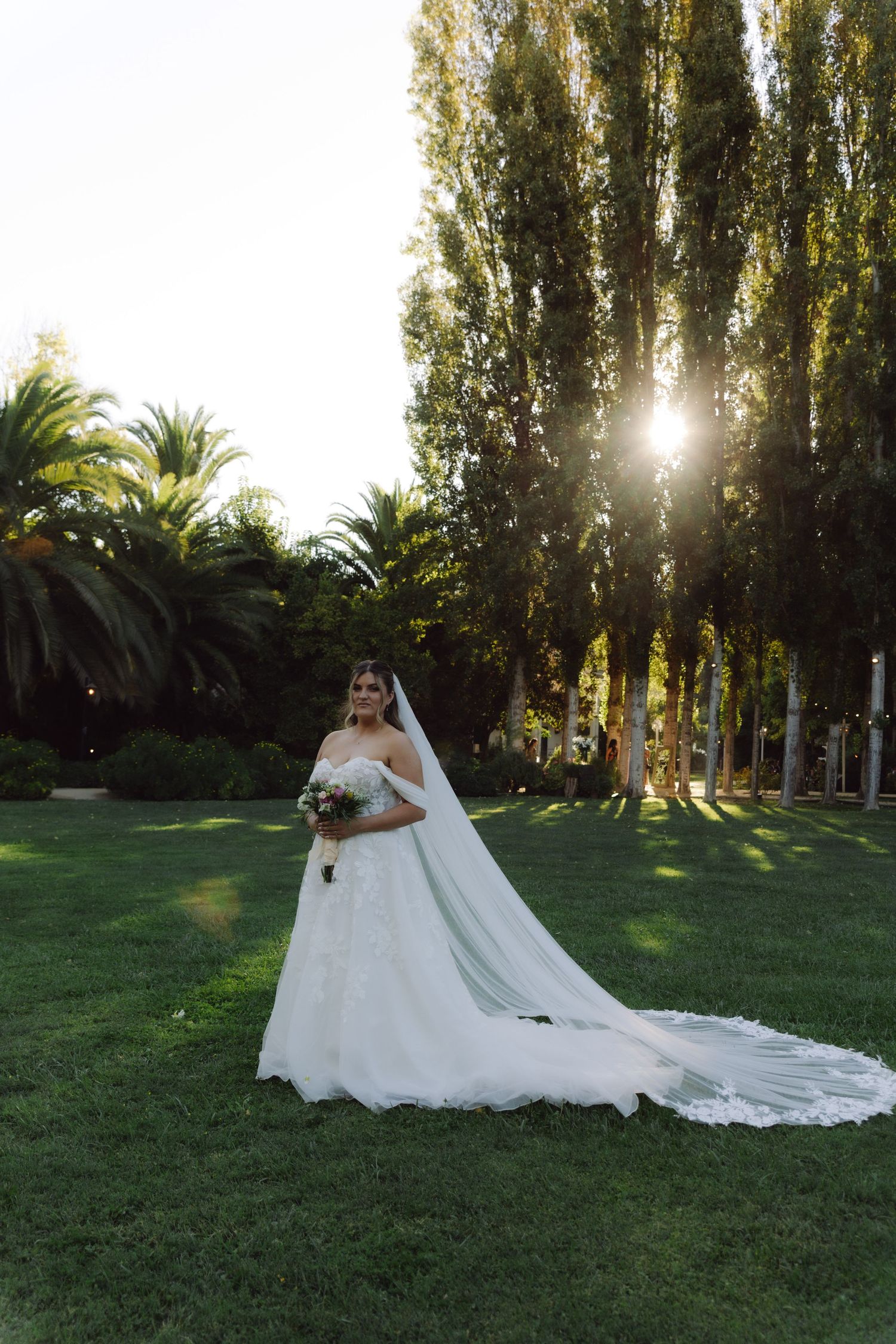 Bride in off-shoulder lace gown with cathedral veil stands in sunlit garden at golden hour.