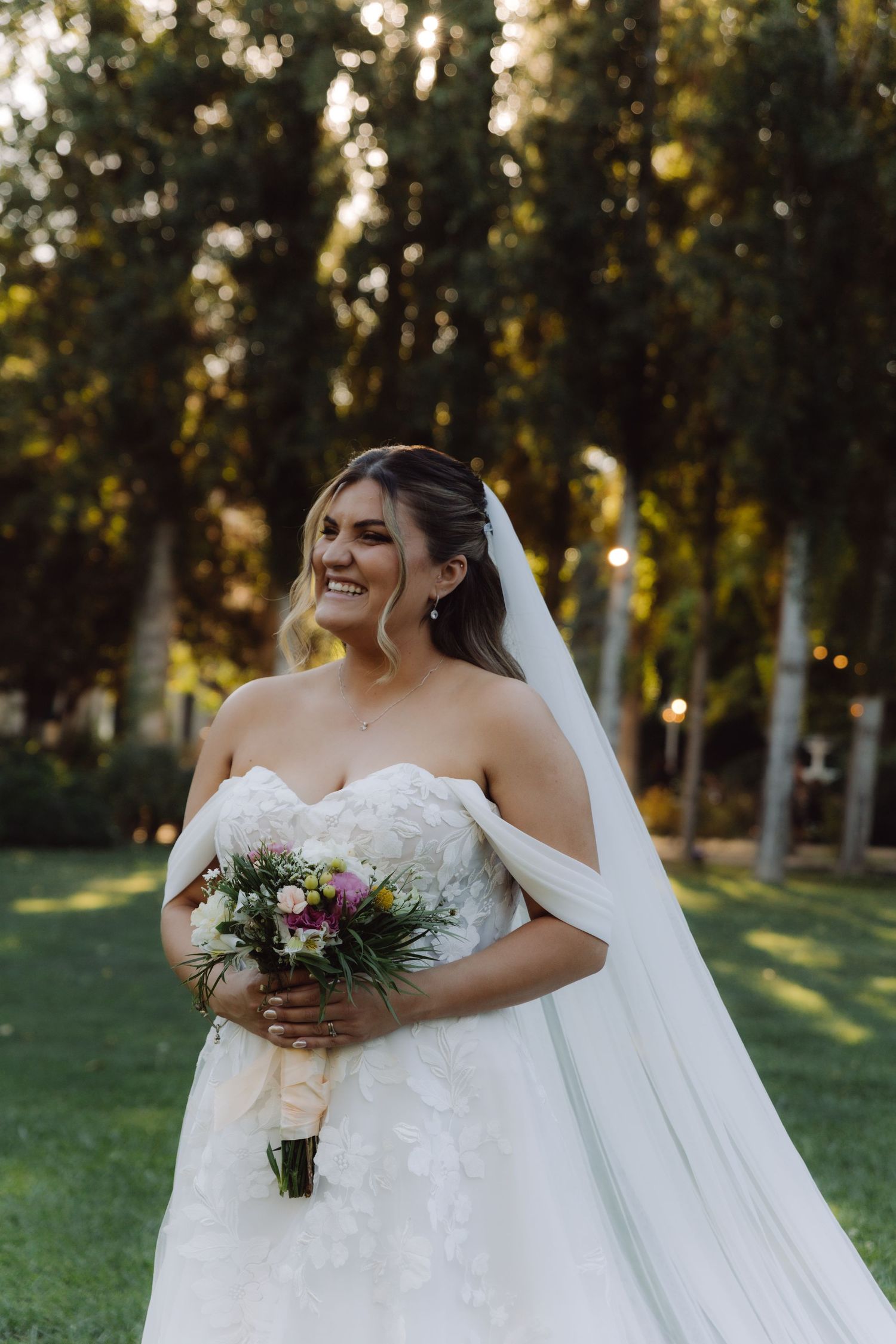 Smiling bride in off-shoulder lace gown holding colorful bouquet outdoors in garden setting.