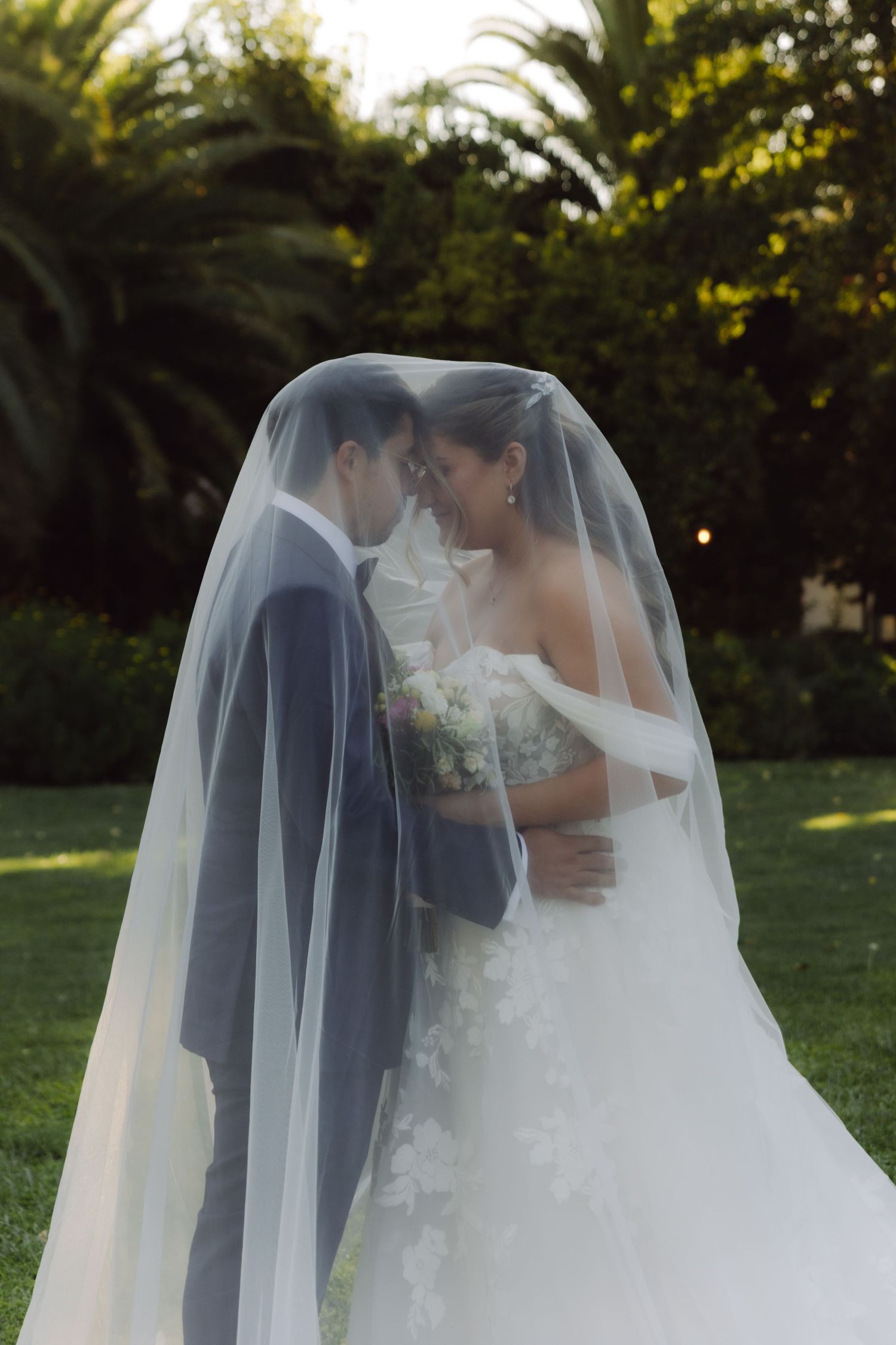 Bride and groom sharing intimate moment together under flowing bridal veil in garden setting.