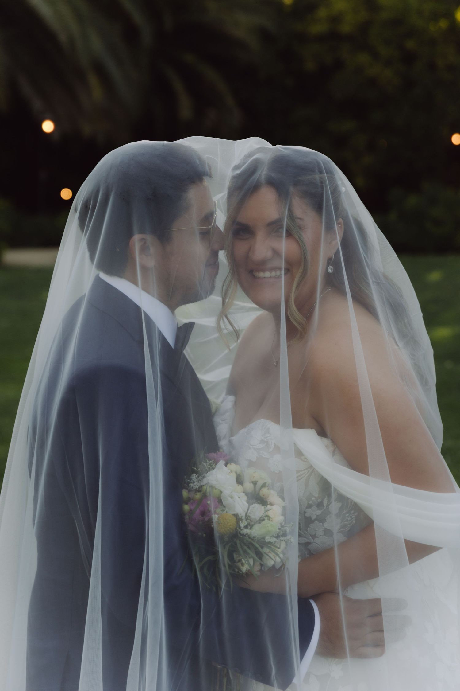 Smiling bride and groom sharing a romantic moment under a sheer veil at dusk.