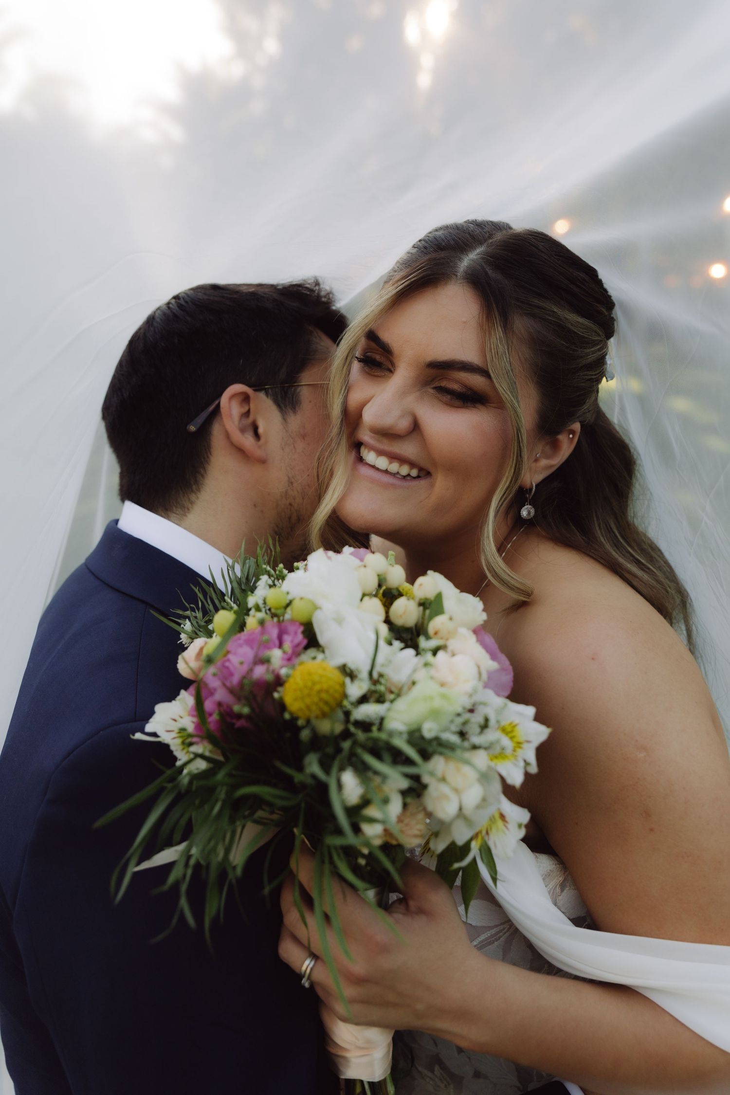Groom kissing bride on cheek as she smiles holding colorful wedding bouquet under veil.