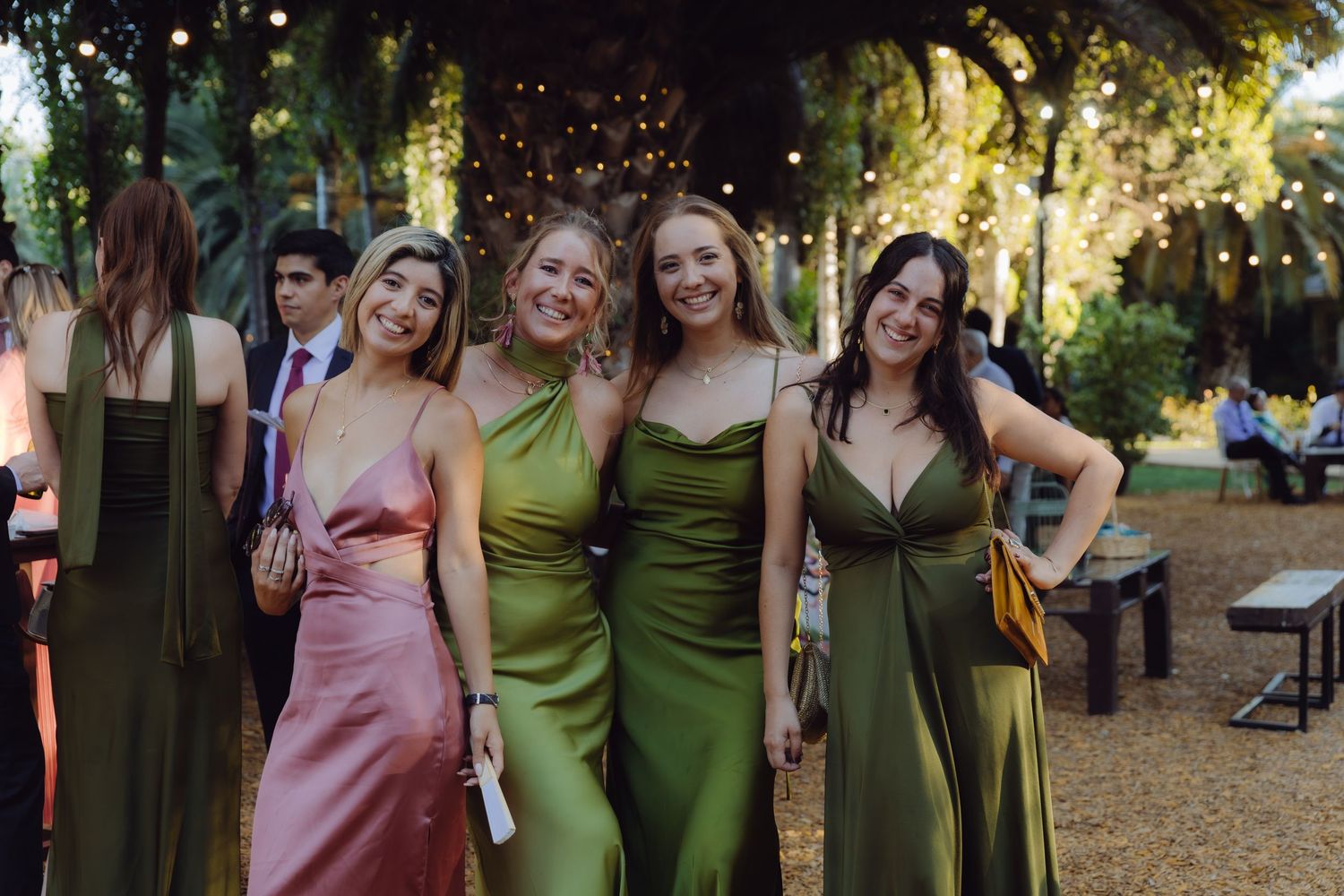 Four smiling bridesmaids in elegant satin green and pink dresses at outdoor wedding celebration.