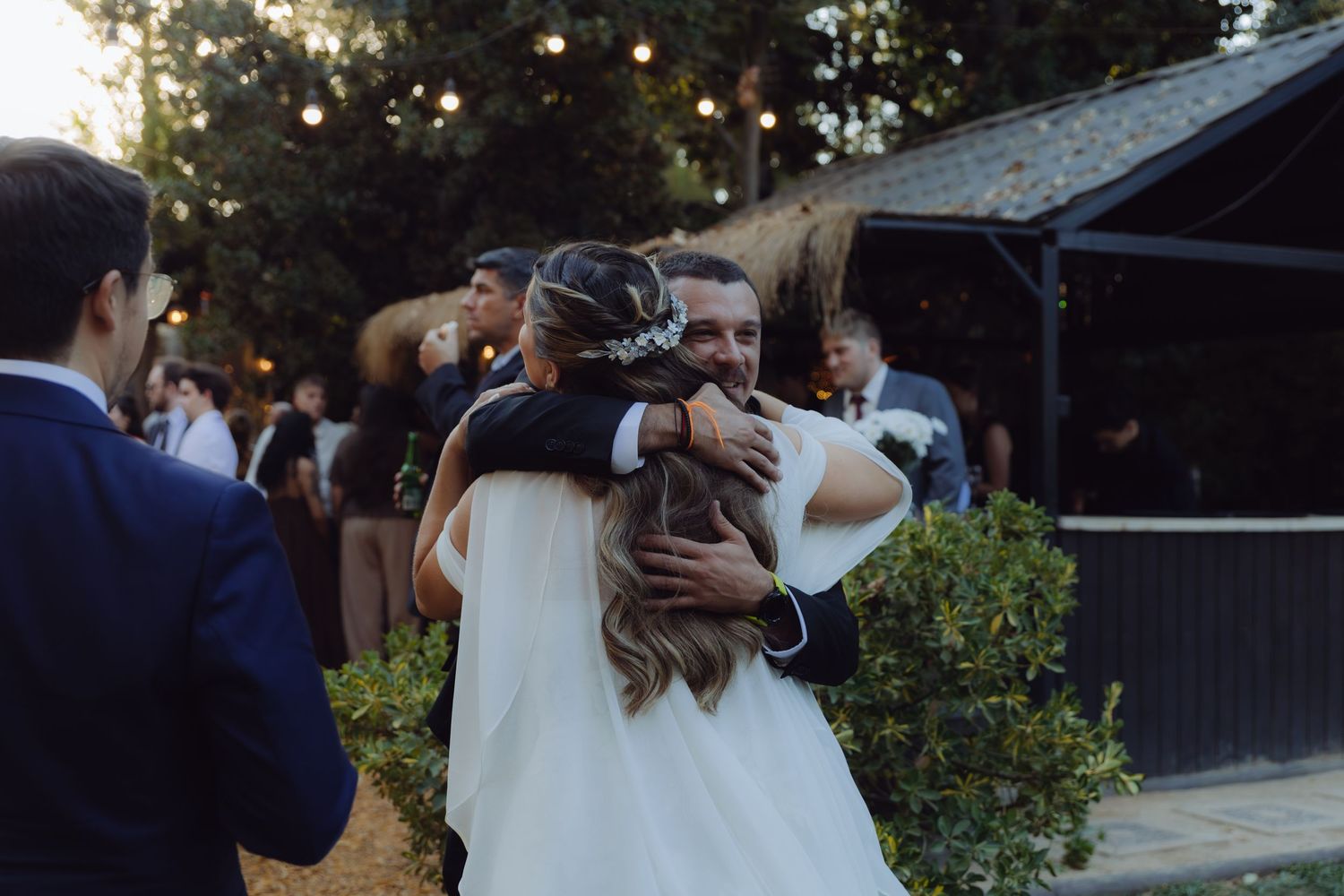 Bride sharing a warm embrace with a guest at outdoor wedding reception under string lights.