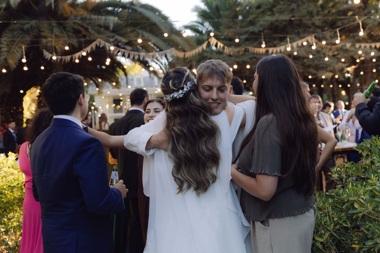 Bride in white gown hugging guests warmly at an outdoor wedding reception with string lights.