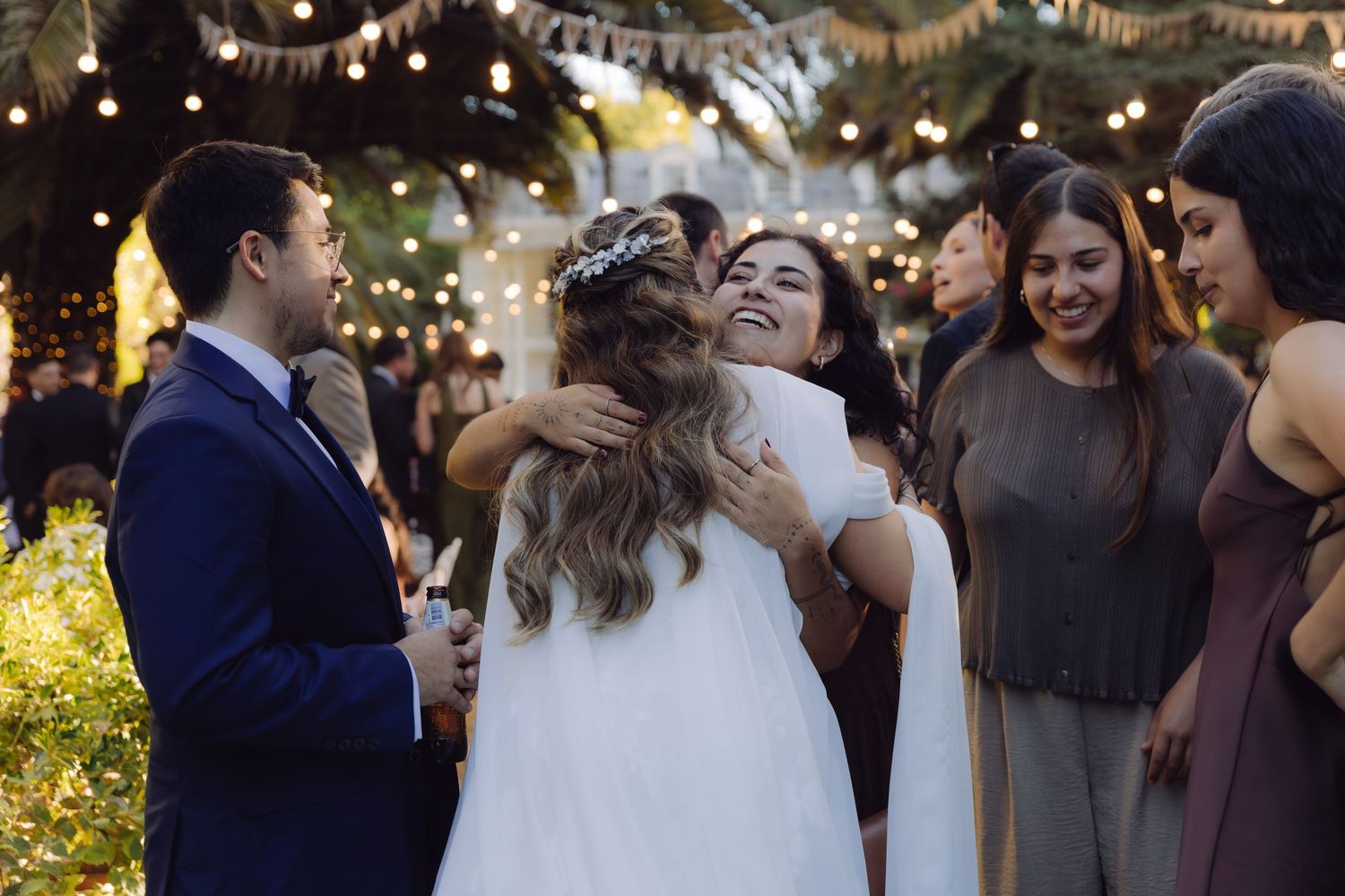 Smiling bride hugging a guest while groom watches at a festive outdoor wedding celebration.