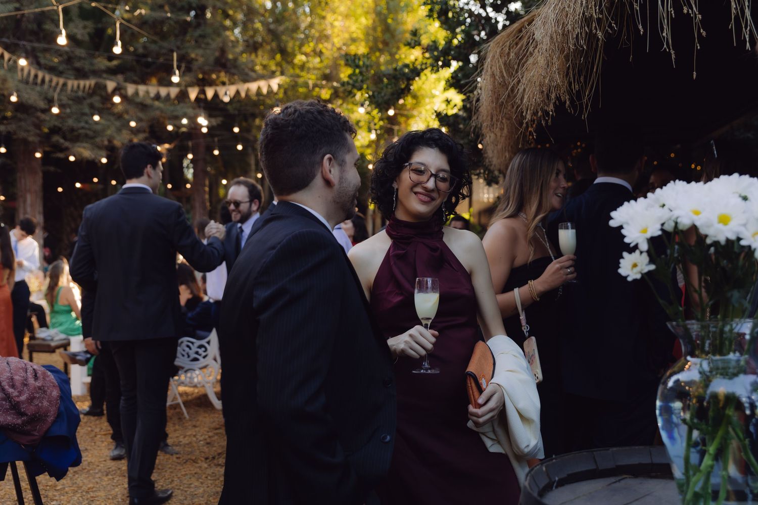 Woman in burgundy dress holding champagne glass chatting with a man at an outdoor evening reception.