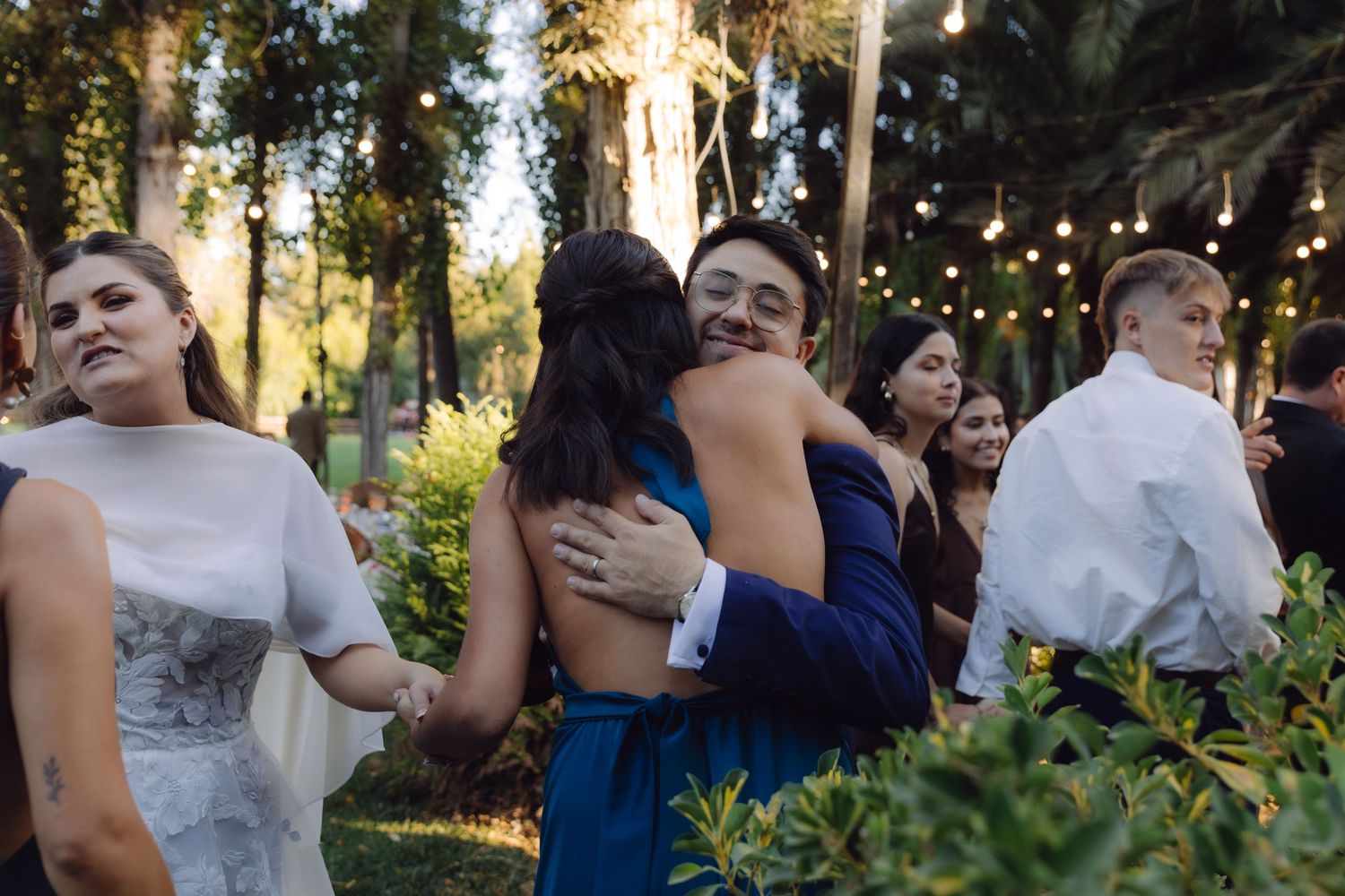 Man in blue suit joyfully hugging a guest at an outdoor wedding reception surrounded by trees.