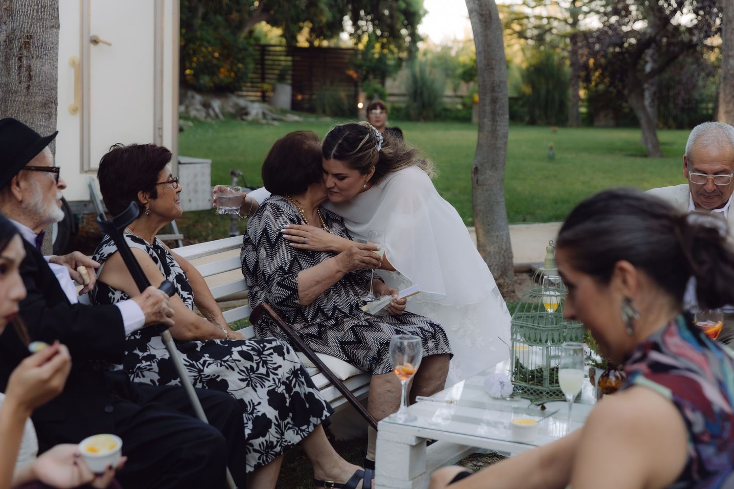 Bride in white dress embracing seated elderly woman at an outdoor wedding reception with guests nearby.