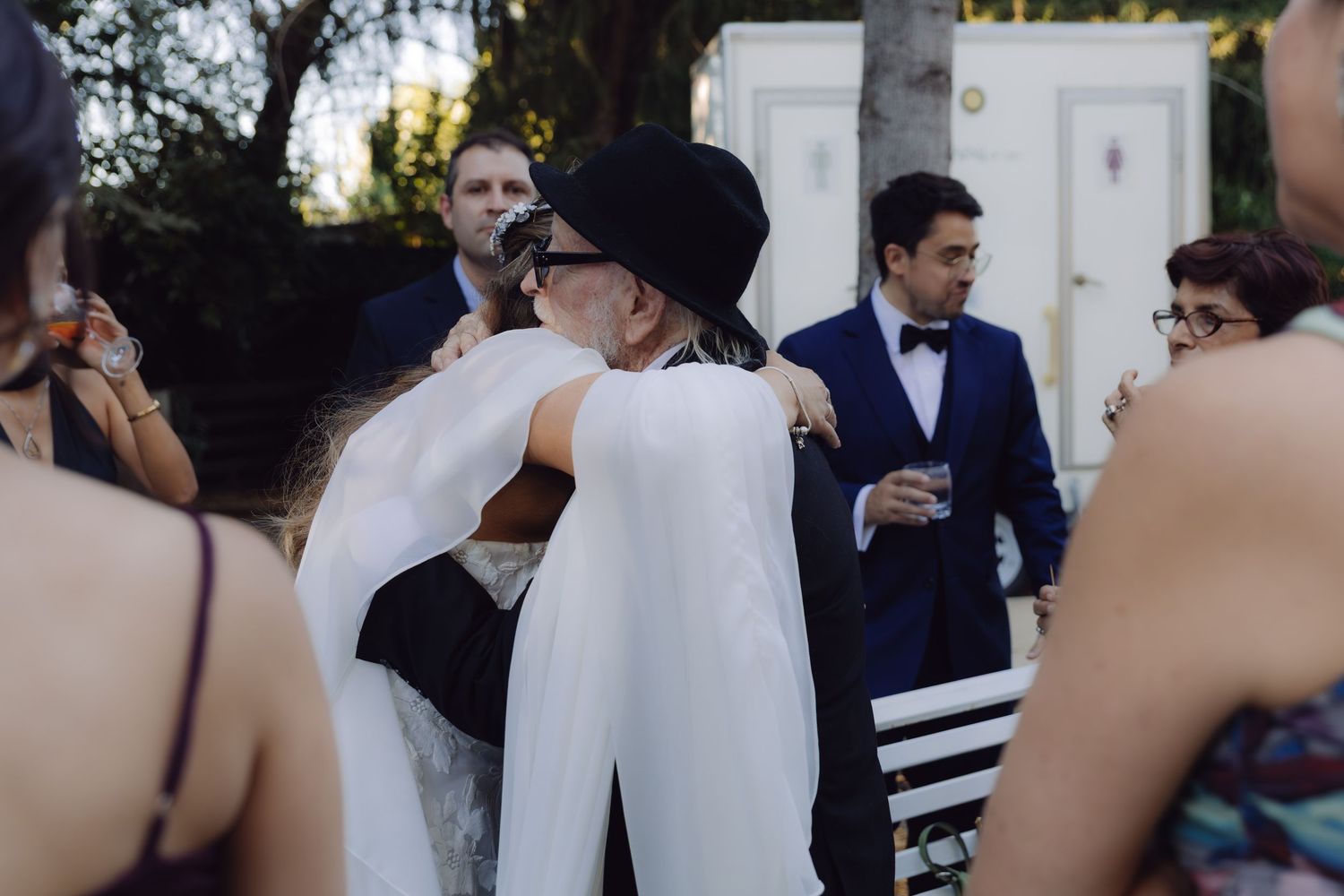 Bride in white cape hugging a guest wearing a black hat at an outdoor wedding ceremony.