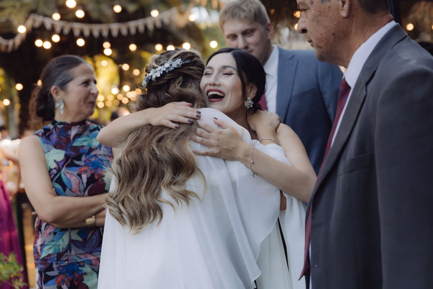 Smiling bride in white dress warmly embracing a guest at an outdoor wedding reception with fairy lights.