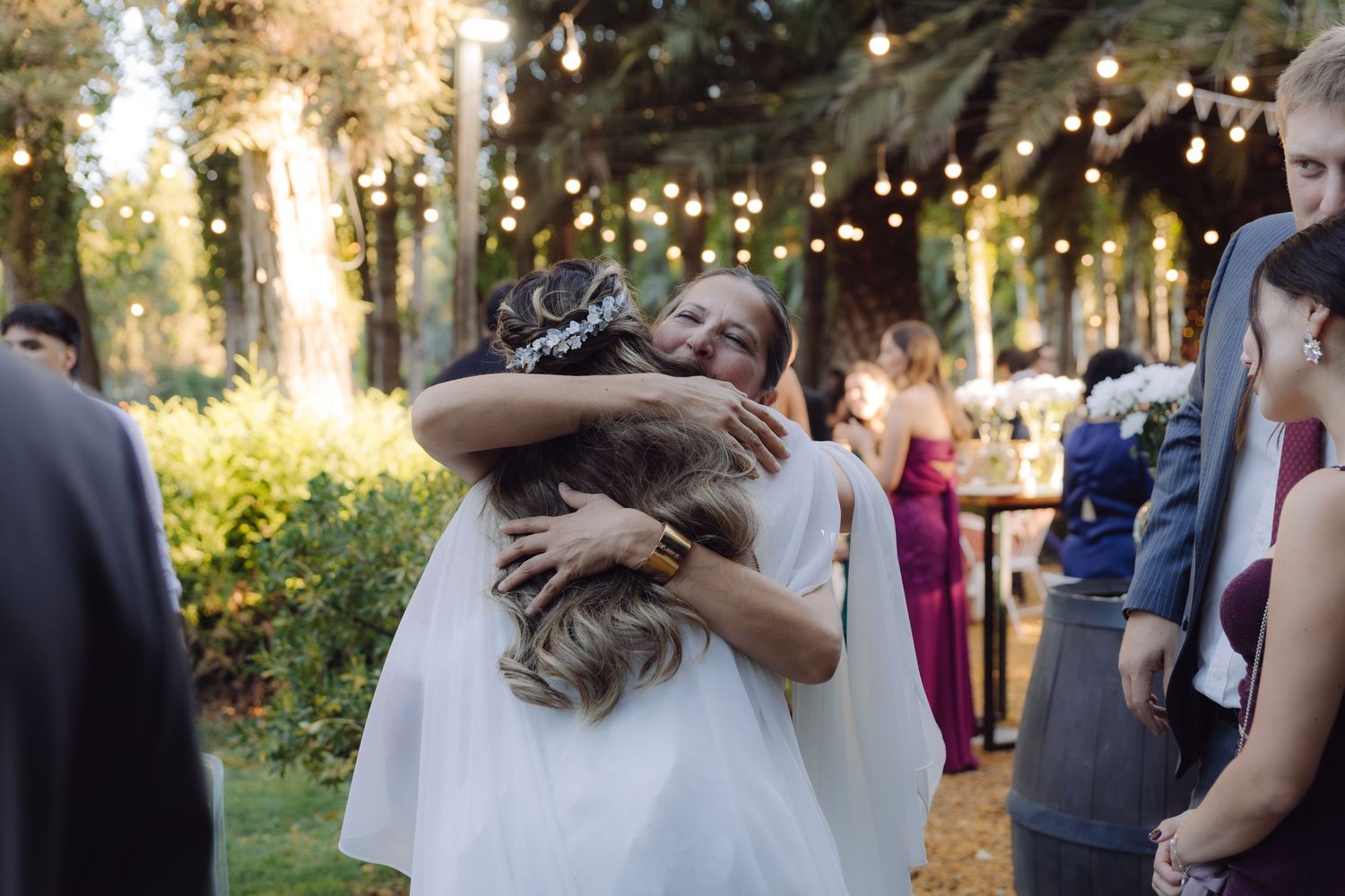 Bride in white dress sharing a warm hug with a guest at a sunlit outdoor wedding reception.