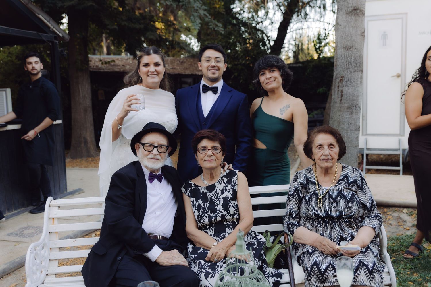 Wedding party group photo with bride, groom in bow tie, and elderly relatives seated outdoors.