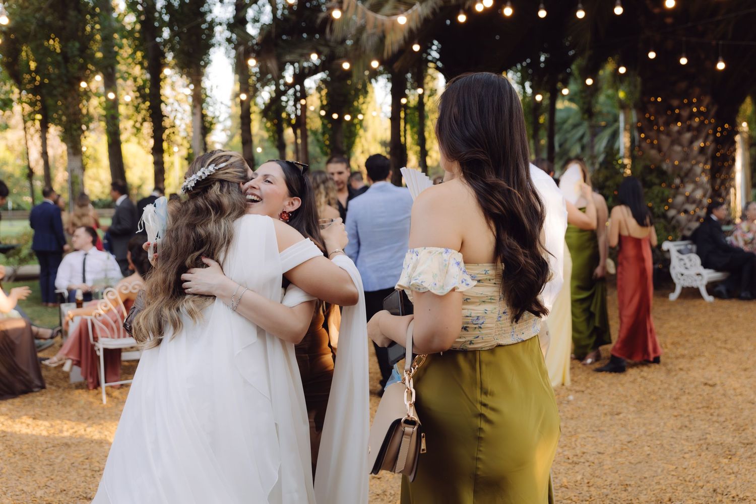 Bride embracing a guest in an olive green dress at an outdoor wedding reception with string lights.