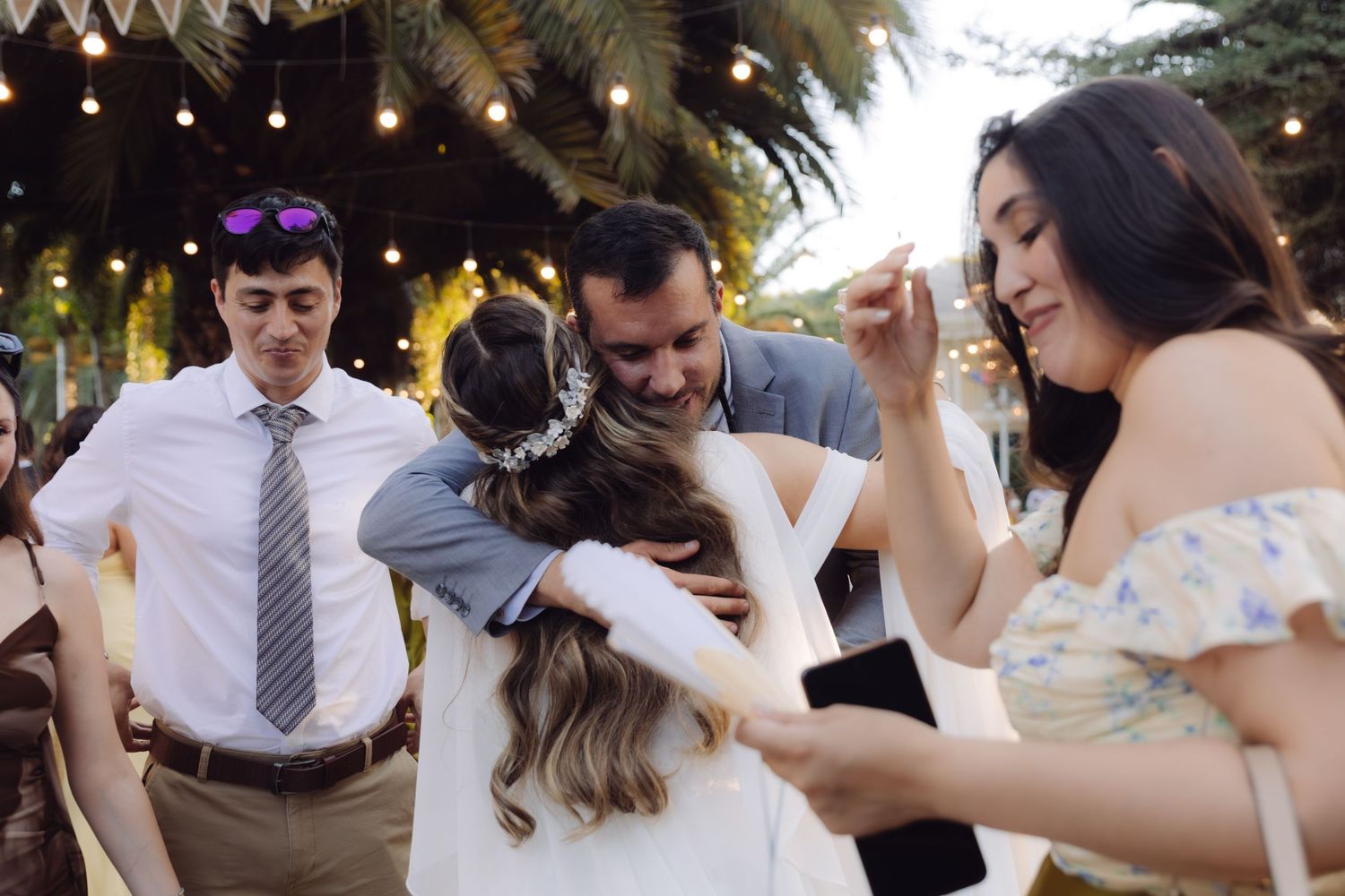 Bride receiving warm hugs from wedding guests at an outdoor evening reception with string lights.