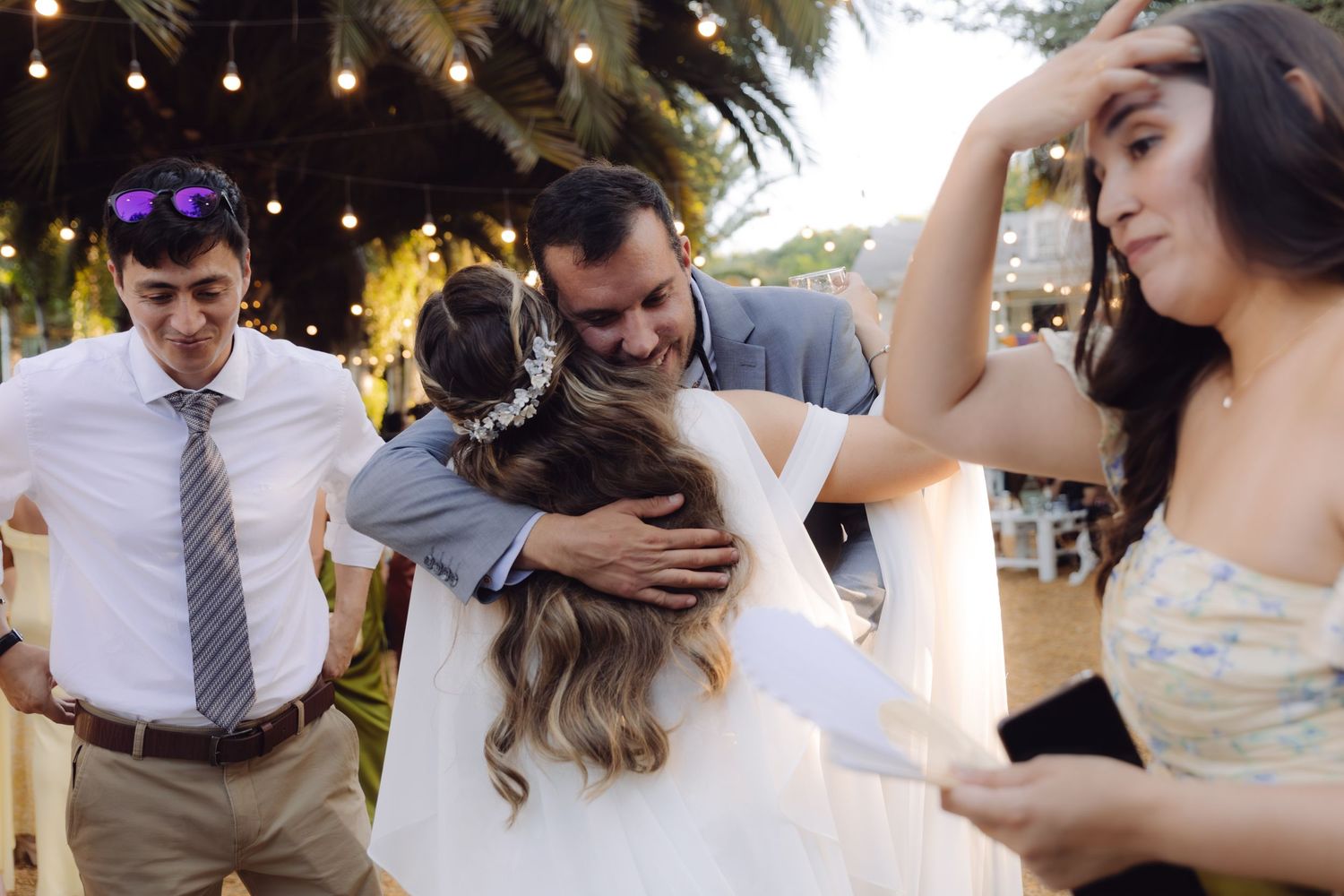 Groom hugging bride surrounded by smiling wedding guests at a festive outdoor evening reception.