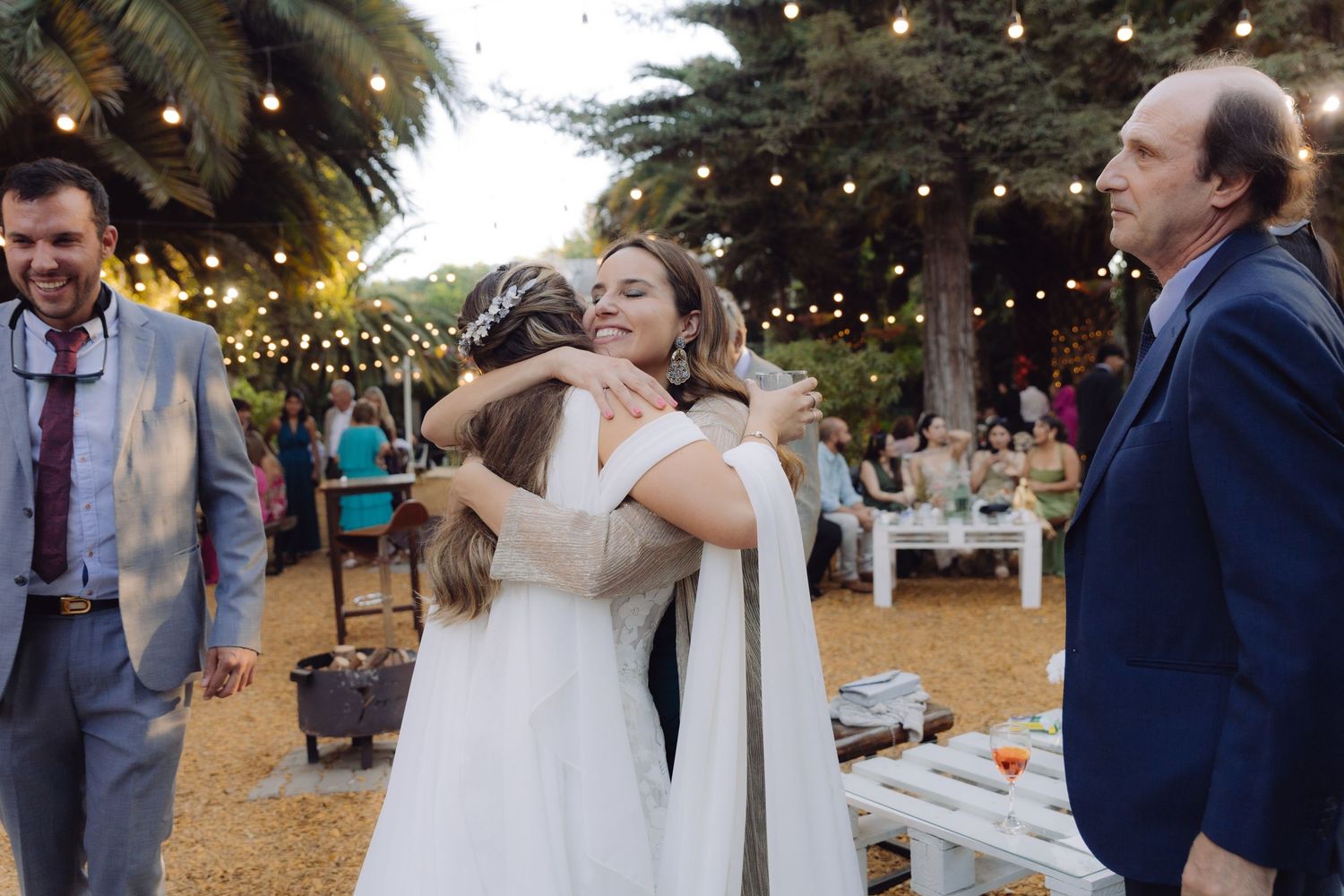 Bride sharing a heartfelt hug with a guest in white at a warmly lit outdoor wedding reception.