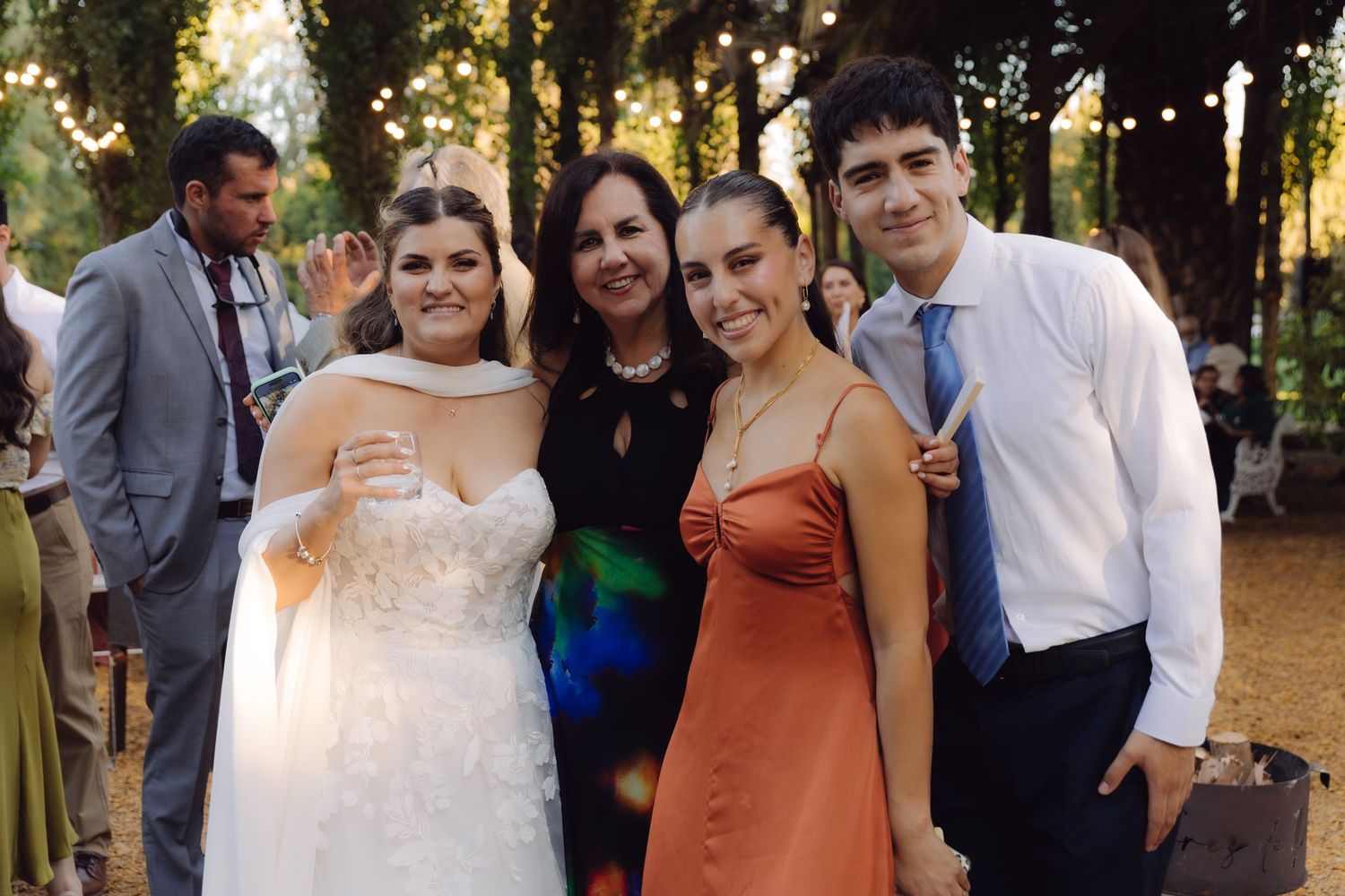 Bride in lace gown posing with three guests including woman in rust dress at outdoor wedding.