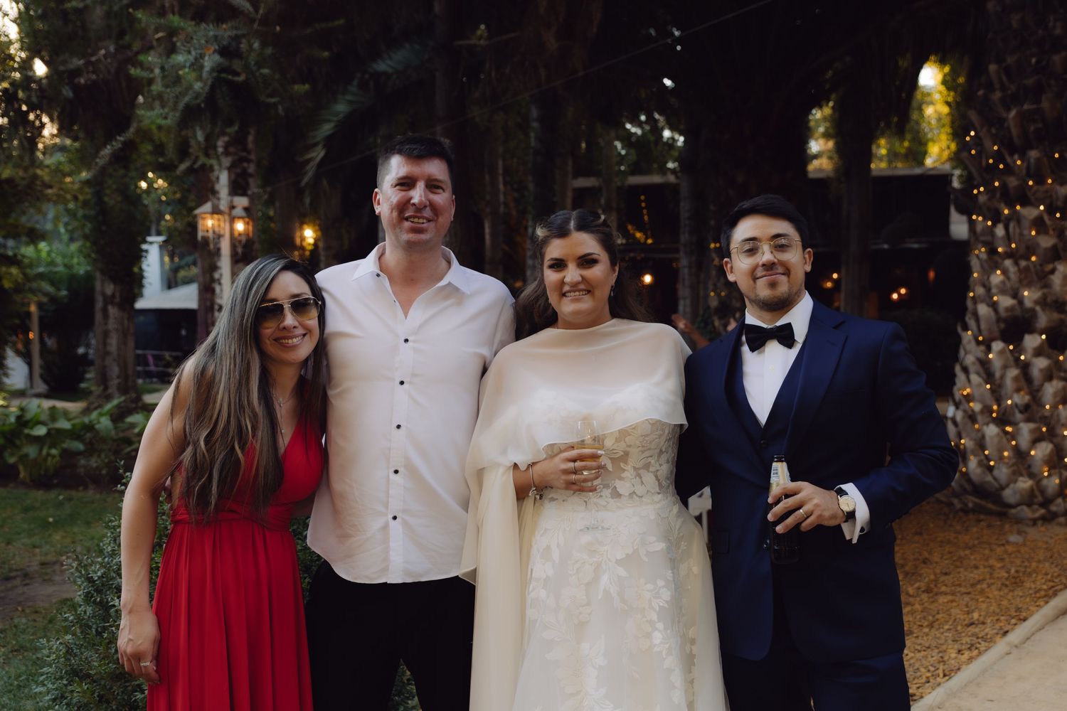 Bride in elegant gown posing with three guests including man in navy tuxedo at outdoor wedding.
