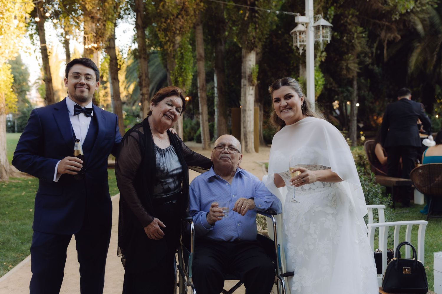 Bride and groom posing with elderly couple in wheelchair at a lush outdoor wedding celebration.