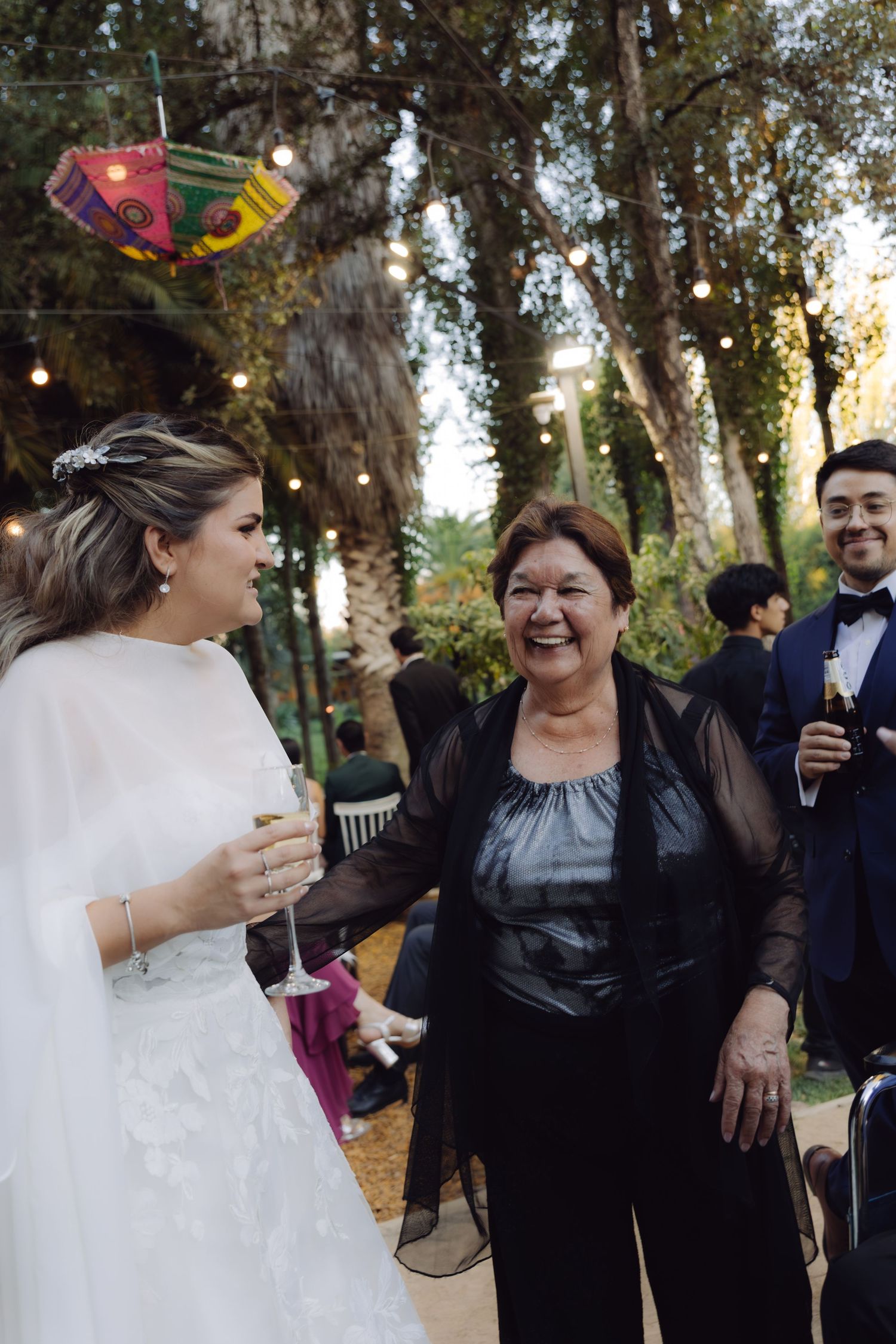 Bride in white gown chatting and smiling with older woman guest at an outdoor evening wedding.