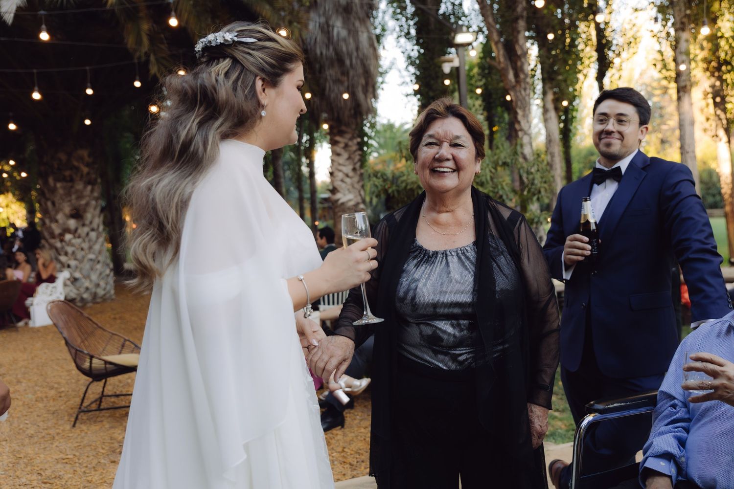 Bride in white gown laughing with older woman in black dress at outdoor wedding reception with string lights.