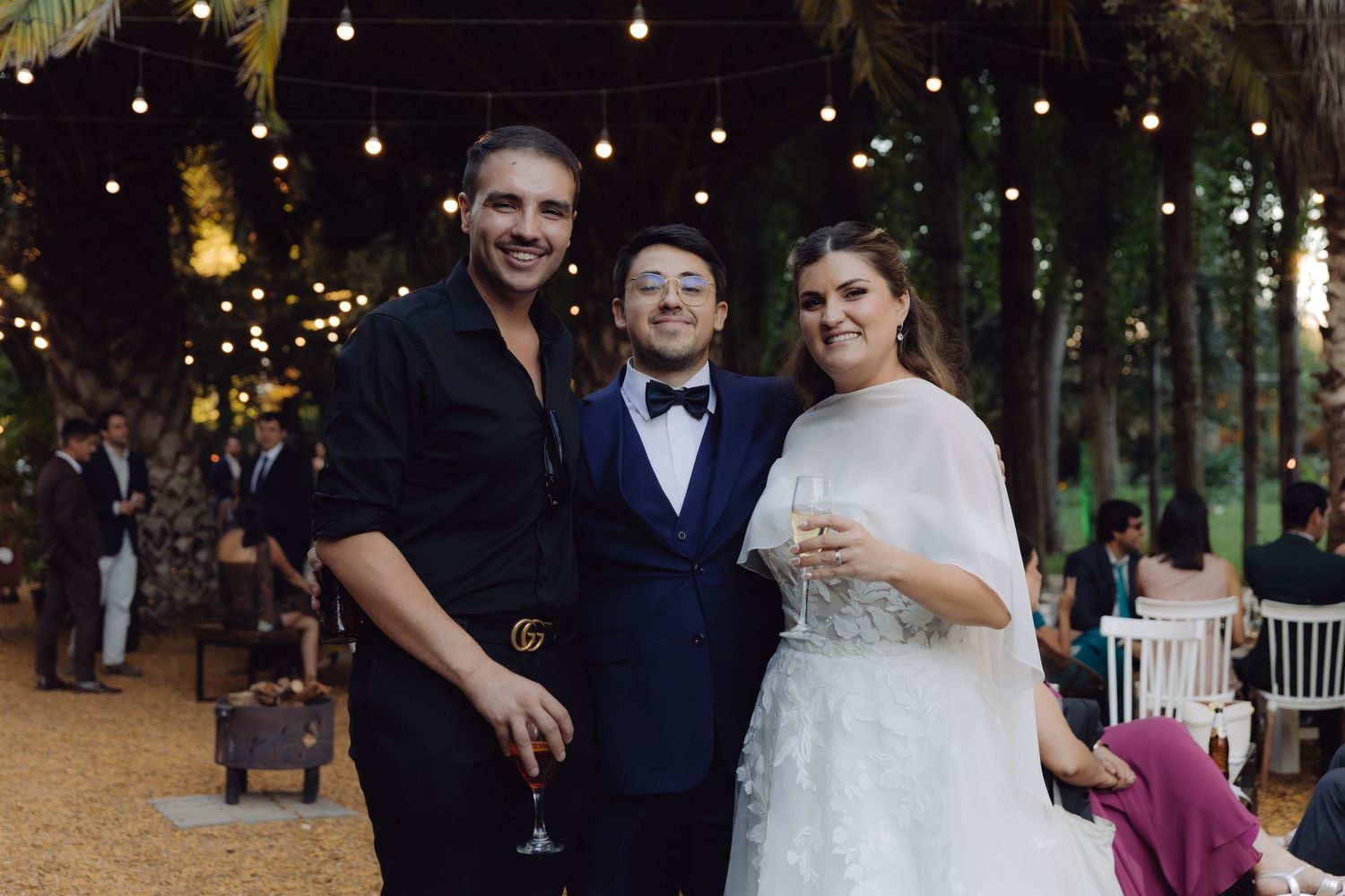 Bride and groom in blue suit posing with guest at elegant outdoor wedding reception with string lights.