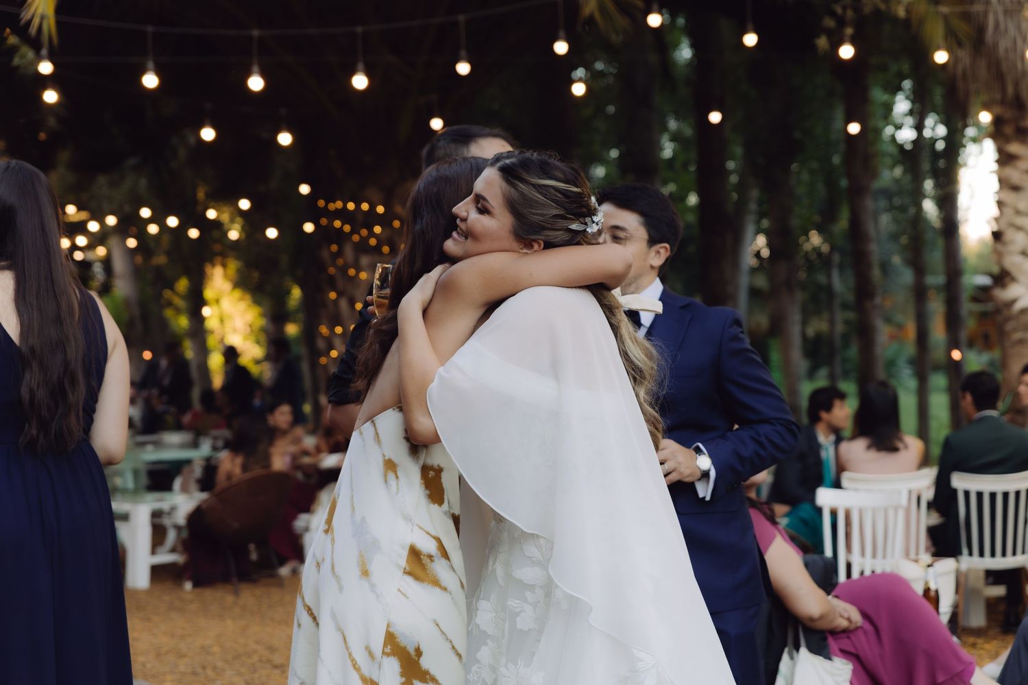 Bride in white wedding gown sharing warm embrace with guest at outdoor evening wedding celebration.