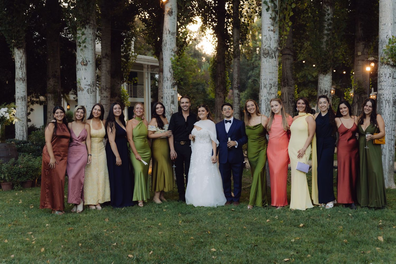 Wedding party posing with bride and groom surrounded by bridesmaids in colorful gowns among birch trees.