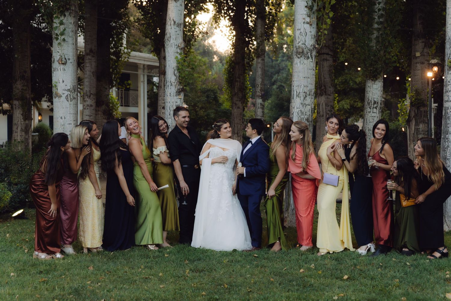 Bride and groom stand with large bridal party in colorful dresses between tall trees at outdoor wedding.