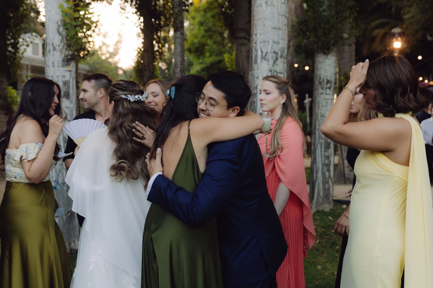 Groom in blue suit hugging bridesmaid in green dress surrounded by wedding guests at outdoor reception.