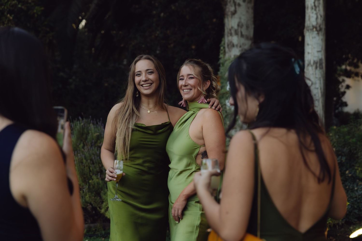 Two women in matching green satin dresses smiling and posing at an elegant outdoor wedding reception.