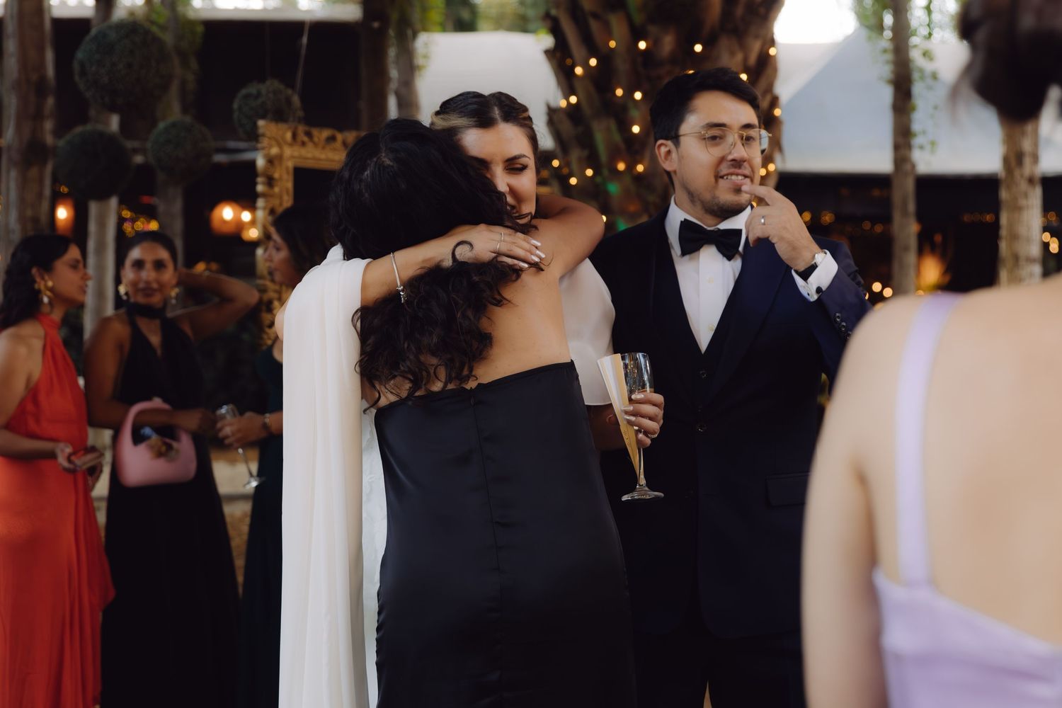 Bride in white cape gown embracing guest in black dress while groom holds champagne at wedding reception.