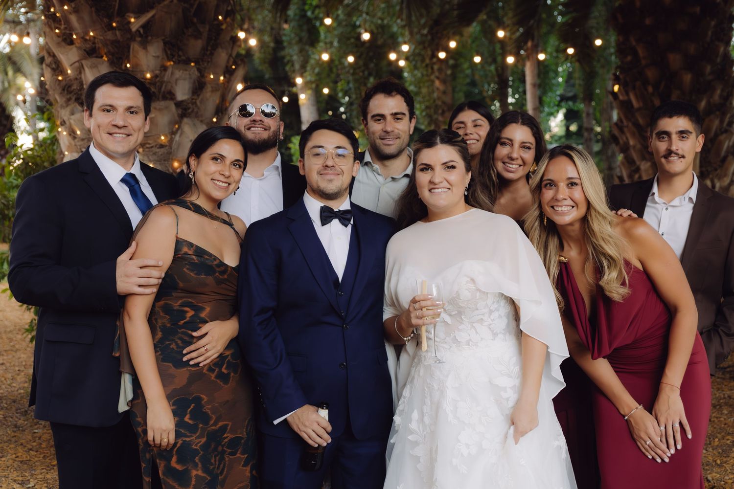 Bride and groom posing with group of smiling friends under string lights at outdoor wedding reception.