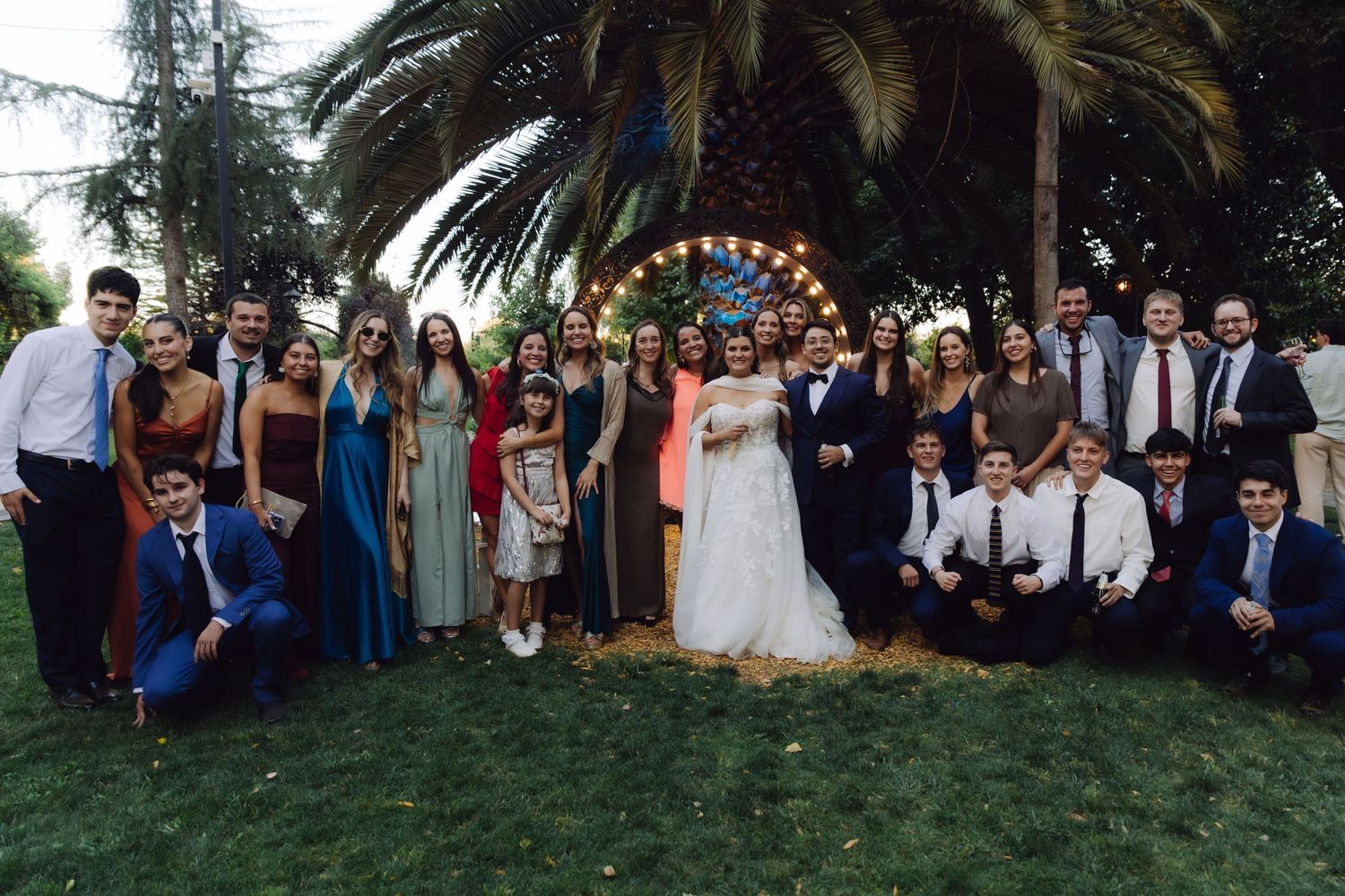 Large wedding group photo with bride in white gown and groom surrounded by guests near decorative arch.