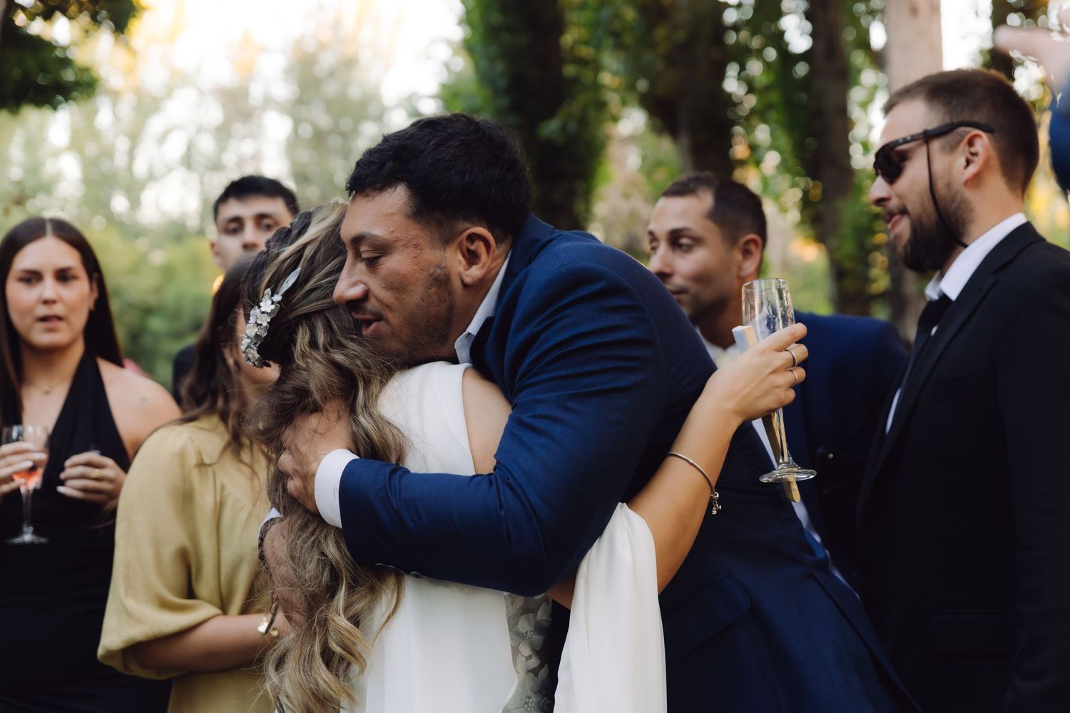 Bride and groom sharing emotional hug with guests holding champagne glasses at outdoor wedding reception.